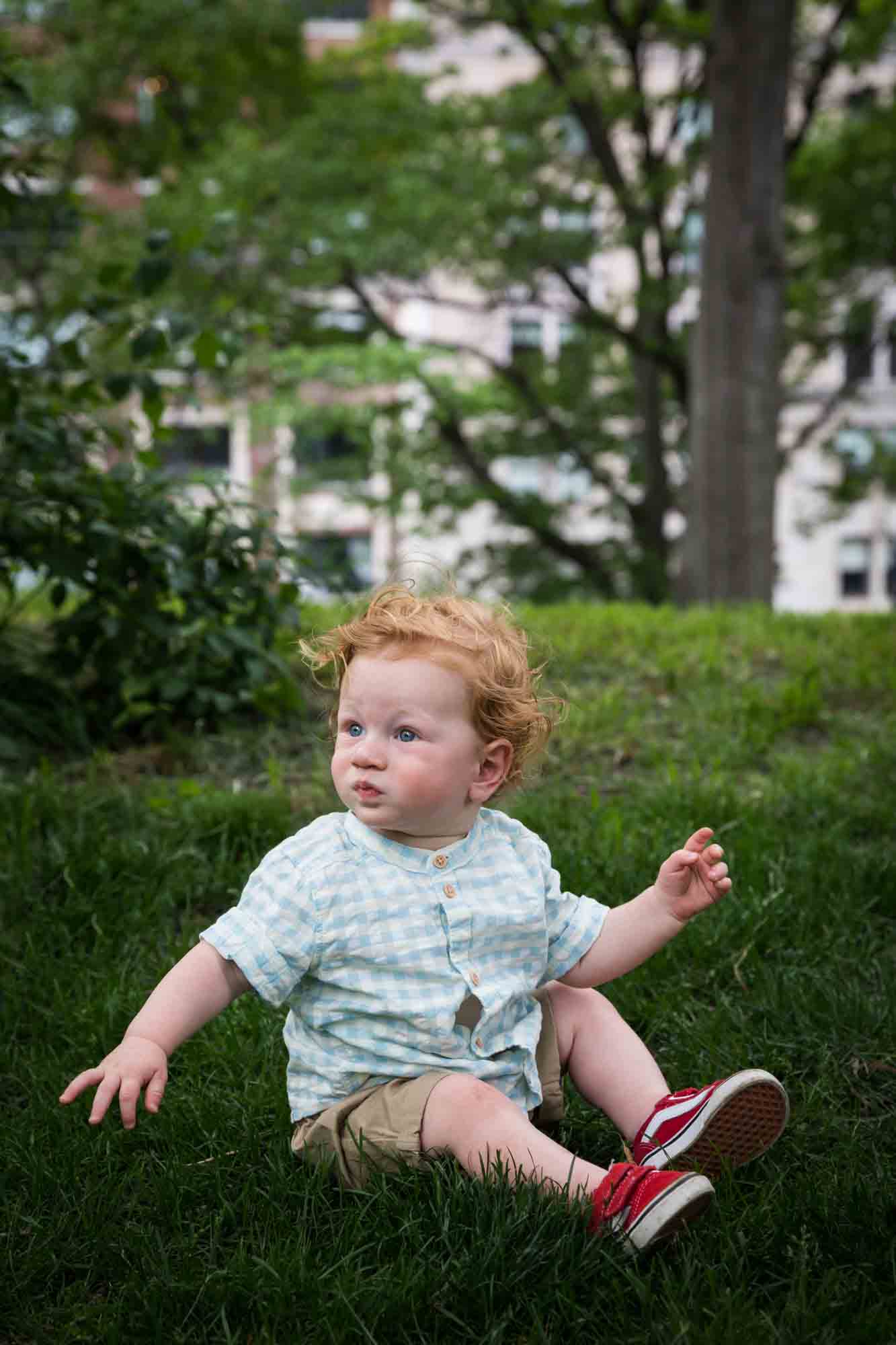 Red-haired baby boy wearing blue and white checked shirt sitting in the grass during a Central Park family portrait session