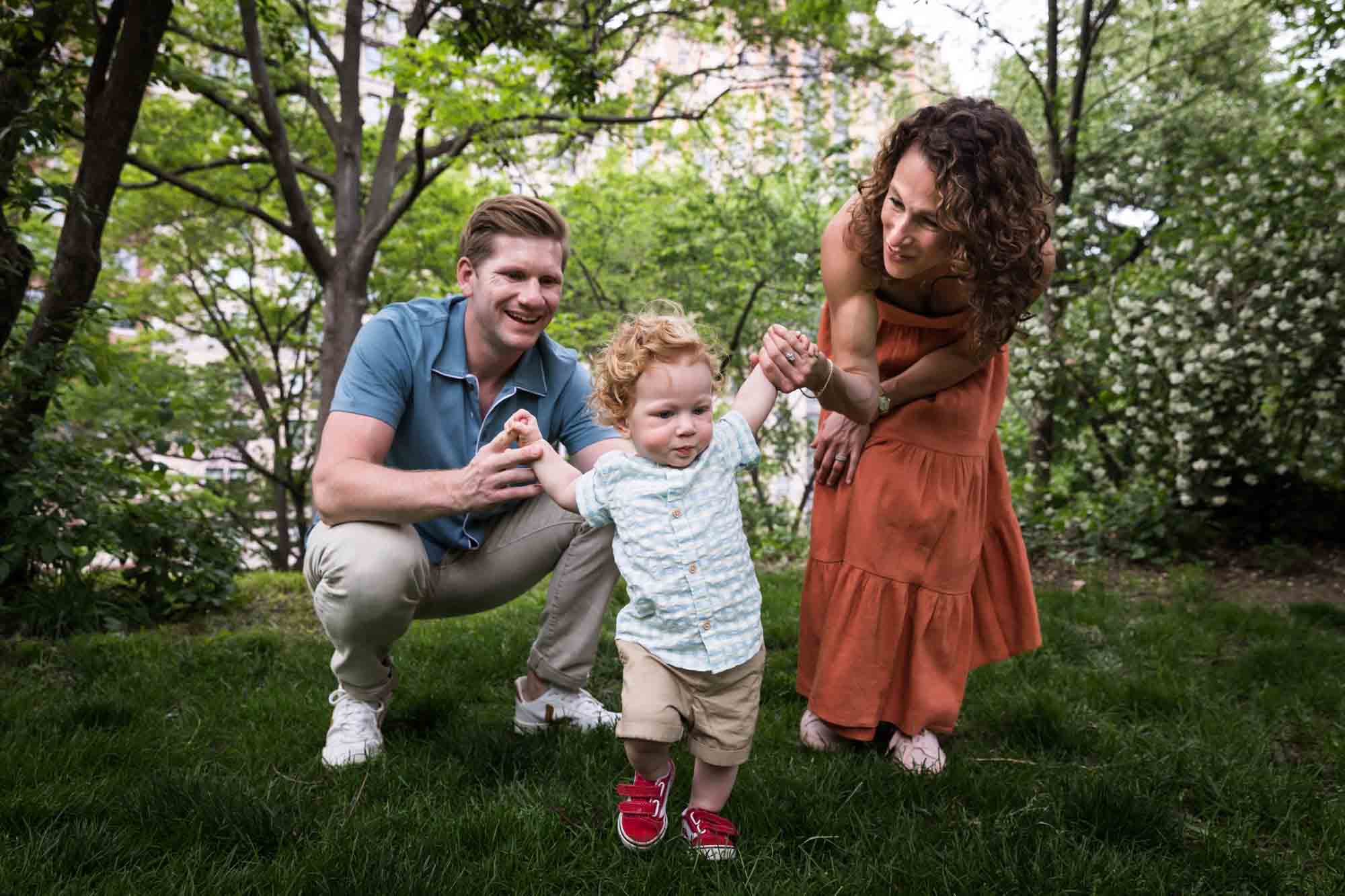 Mother and father helping red-haired baby boy walk in grass during a Central Park family portrait session