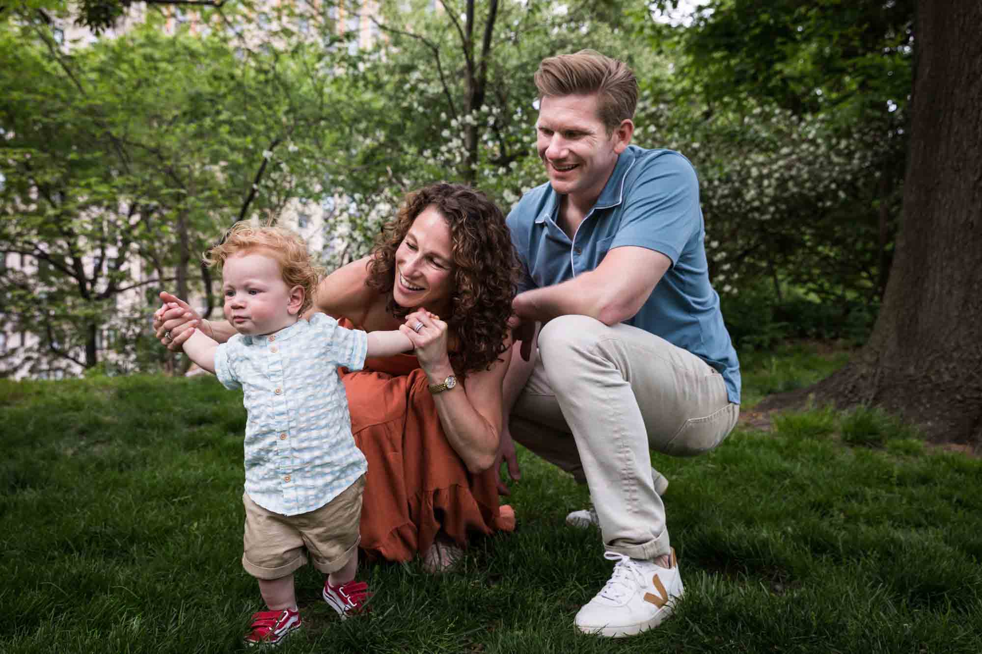 Mother and father helping red-haired baby boy walk in grass during a Central Park family portrait session