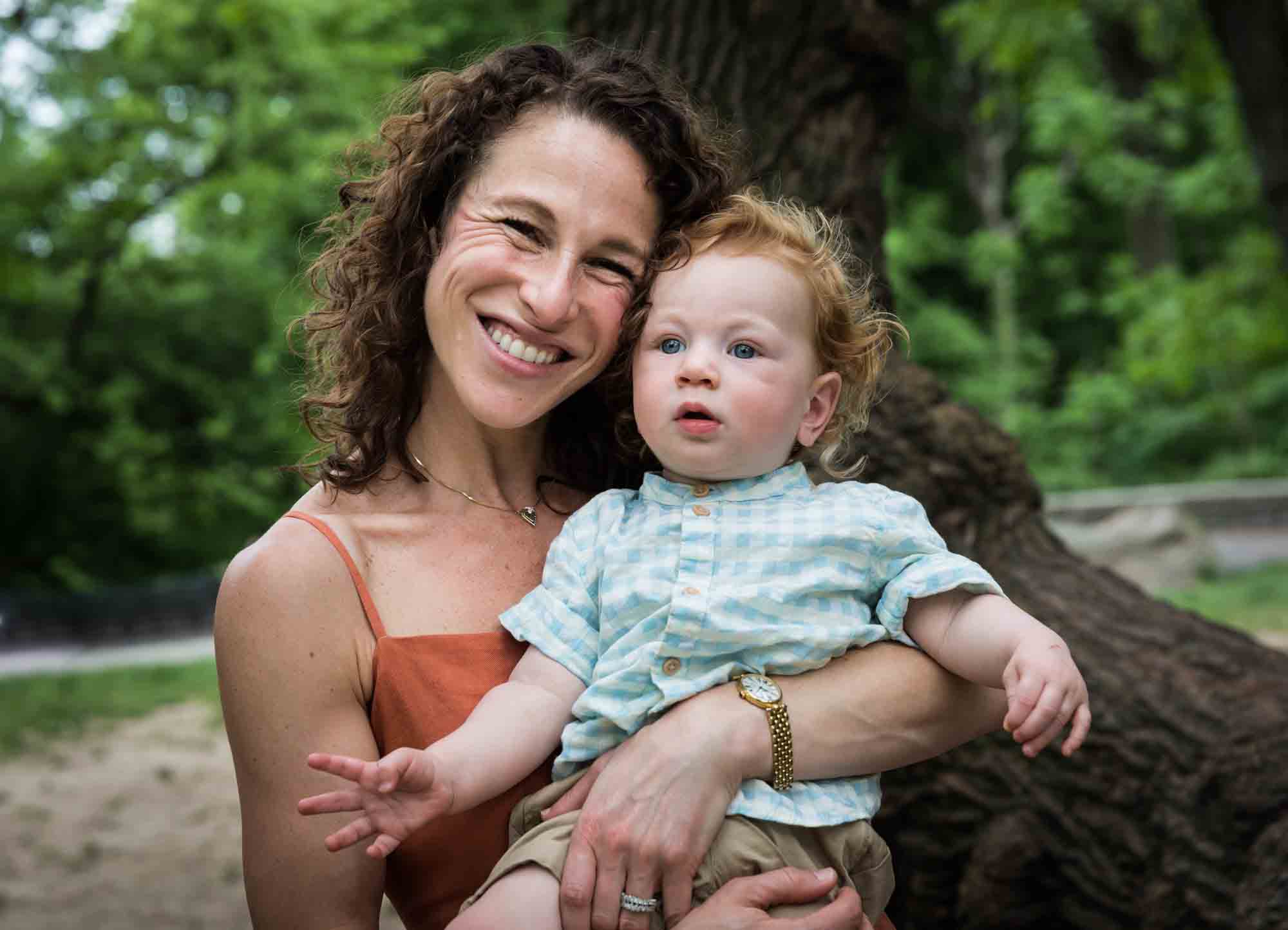 Mother holding red-haired baby boy in the air in front of trees during a Central Park family portrait session