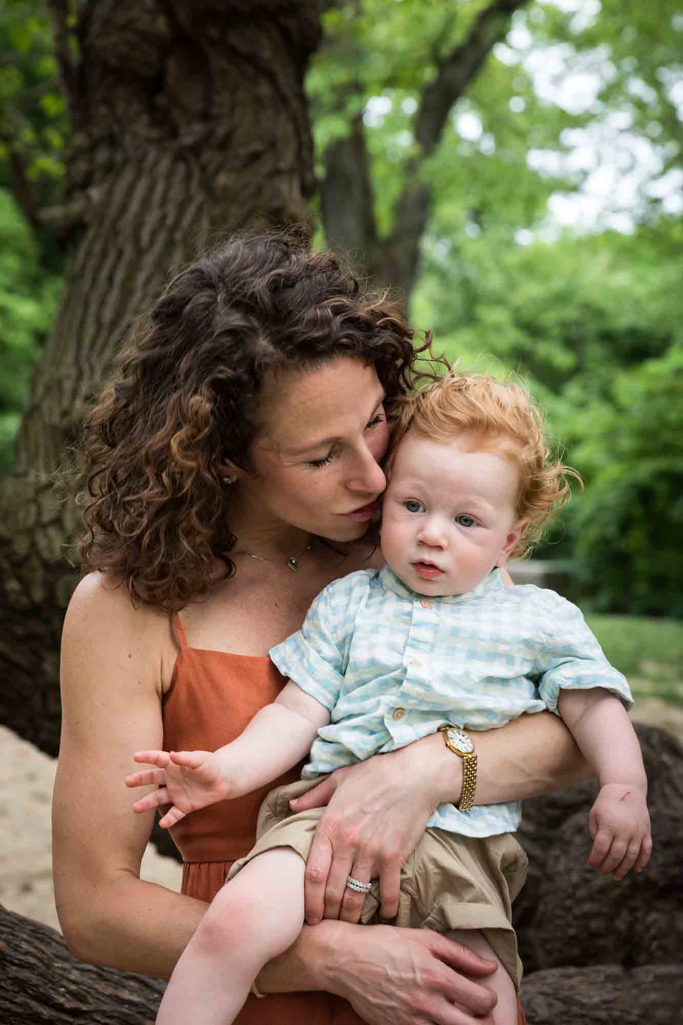Mother holding red-haired baby boy in the air in front of trees during a Central Park family portrait session
