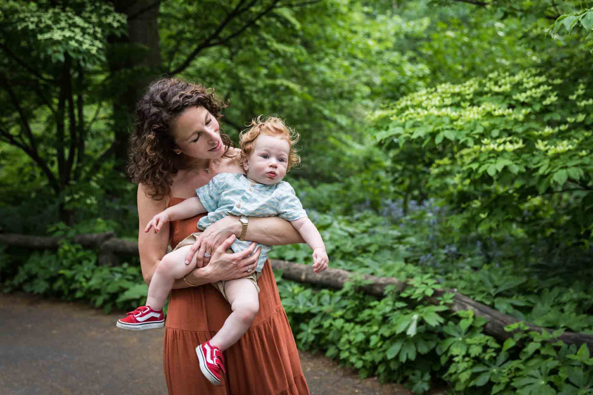 Mother holding red-haired baby boy in the air in front of trees during a Central Park family portrait session