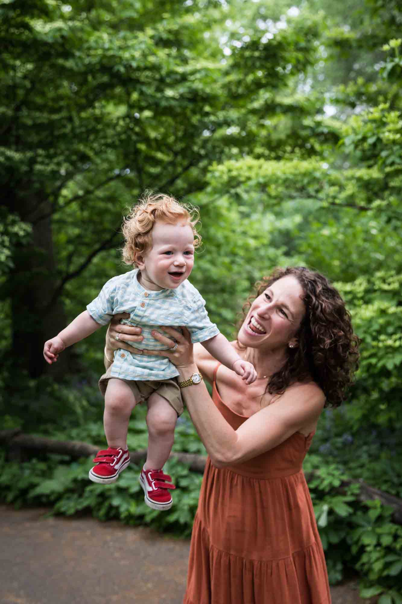 Mother holding red-haired baby boy in the air in front of trees during a Central Park family portrait session