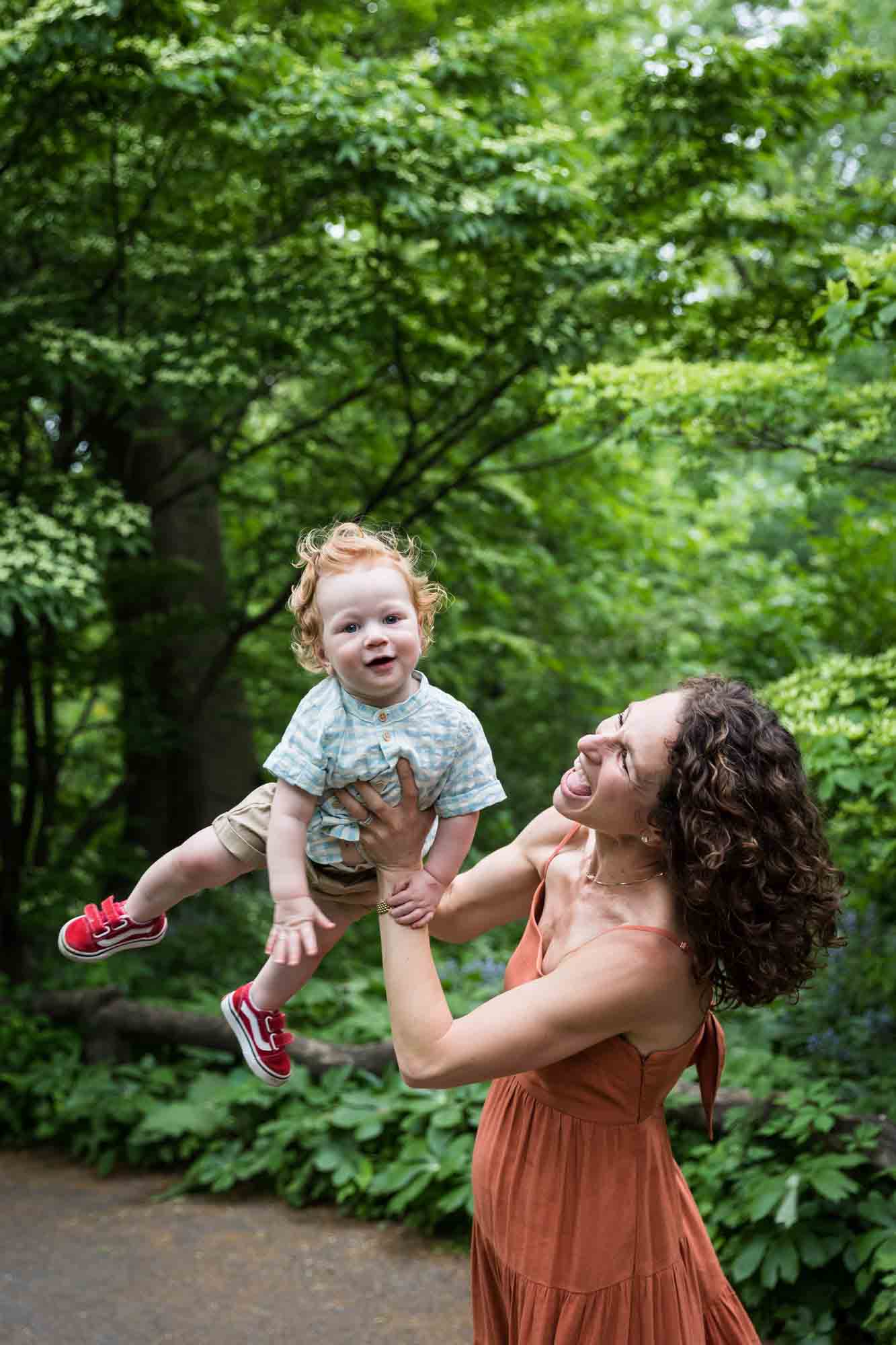 Mother holding red-haired baby boy in the air in front of trees during a Central Park family portrait session