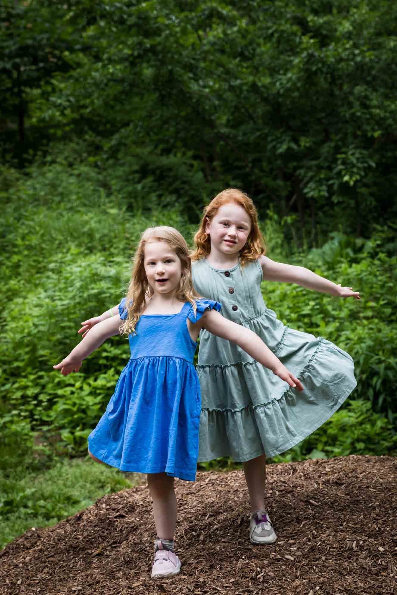 Two little girls wearing blue dresses dancing on mulch pile in front of trees during a Central Park family portrait session
