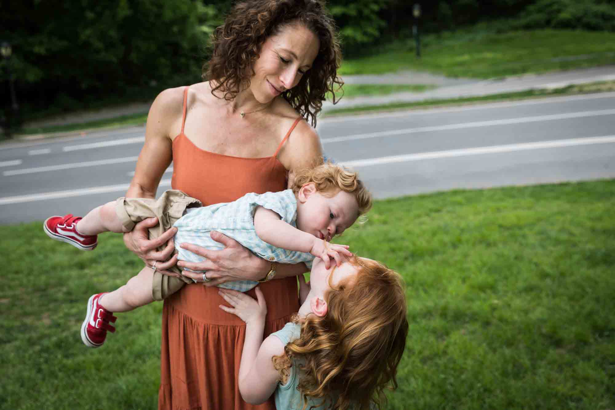 Mother holding baby boy playing with sister's face in grass during a Central Park family portrait session