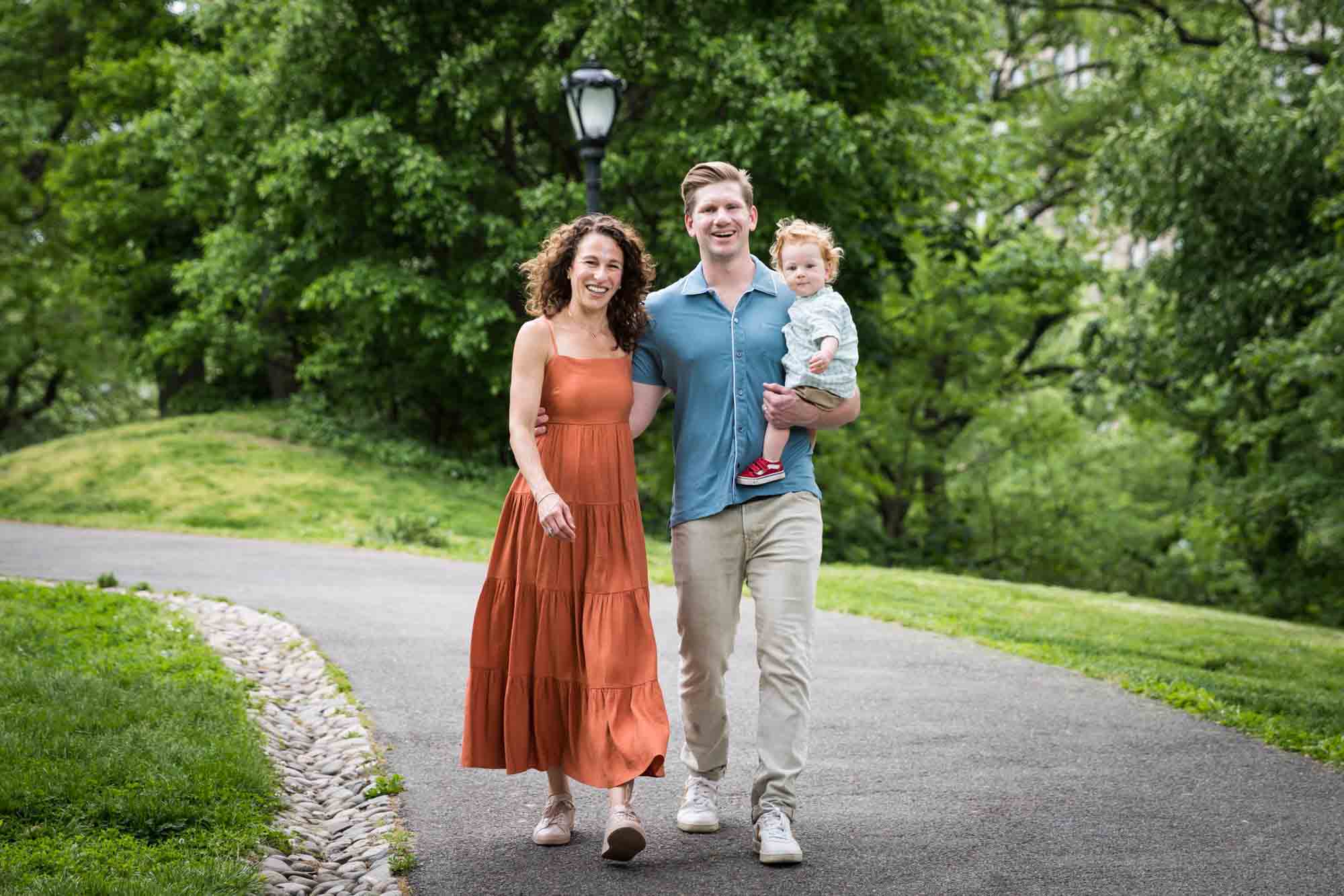 Mother and father holding red-haired little boy walking up pathway in front of trees during a Central Park family portrait session