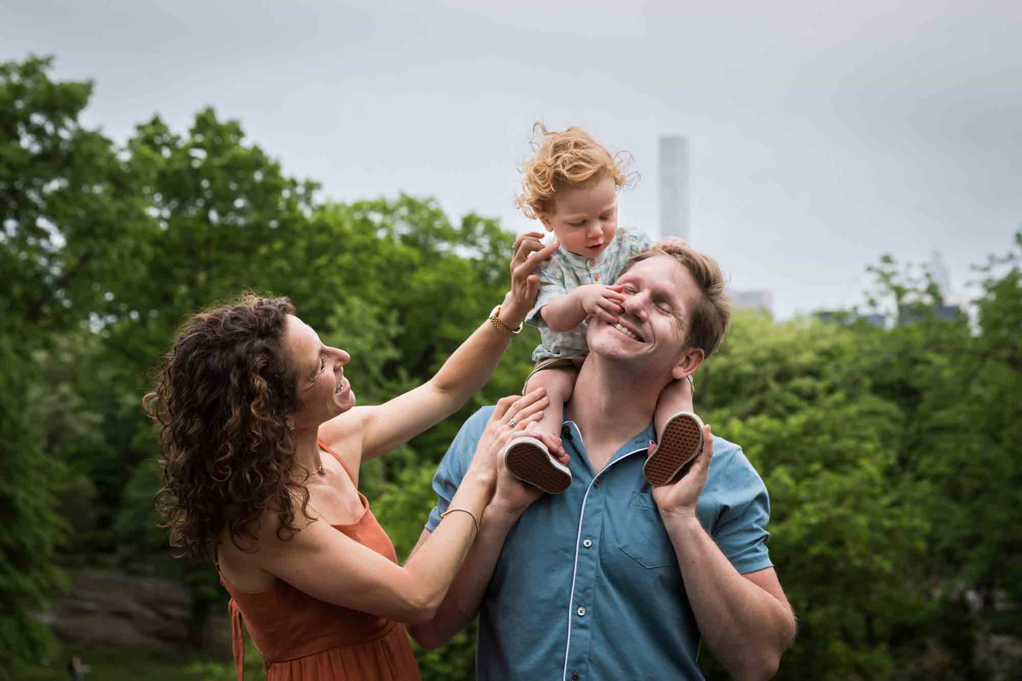 Mother and father looking back at red-haired baby boy held on father's shoulders during a Central Park family portrait session
