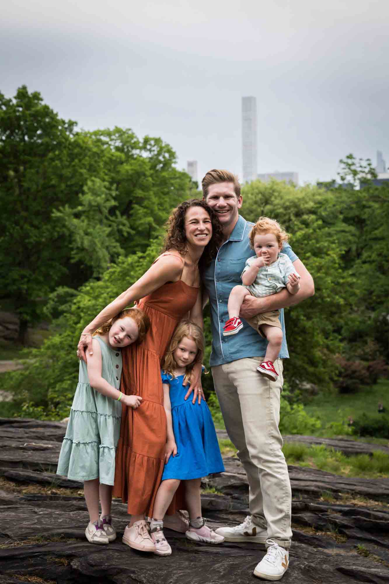 Mother, father and three children standing on rock in front of trees and NYC skyline during a Central Park family portrait session