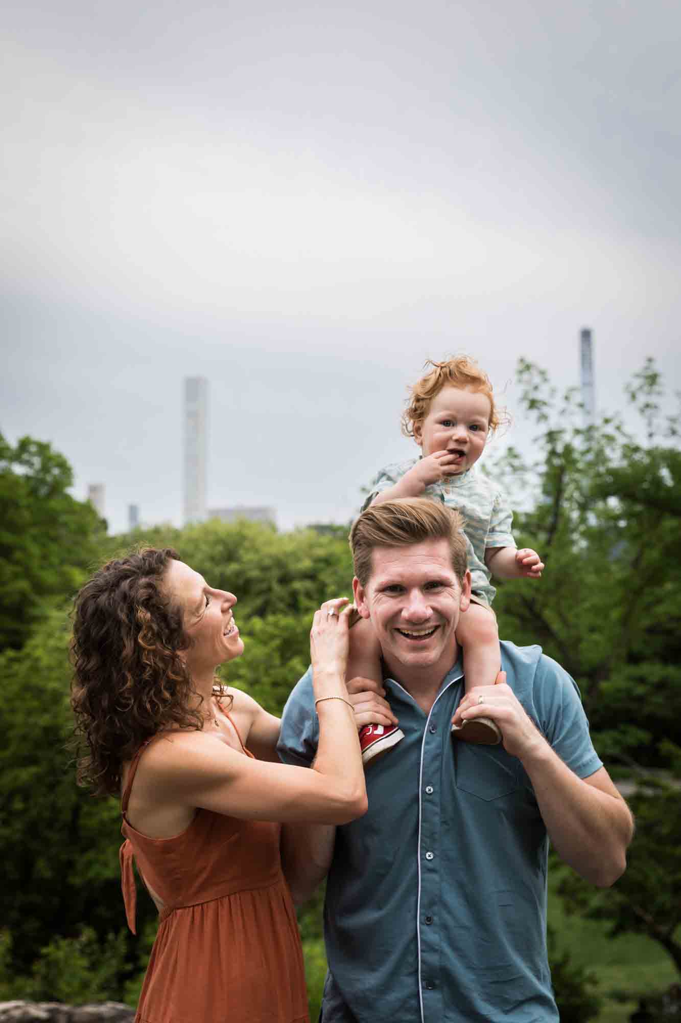 Mother and father playing with red-haired baby boy held on father's shoulders during a Central Park family portrait session