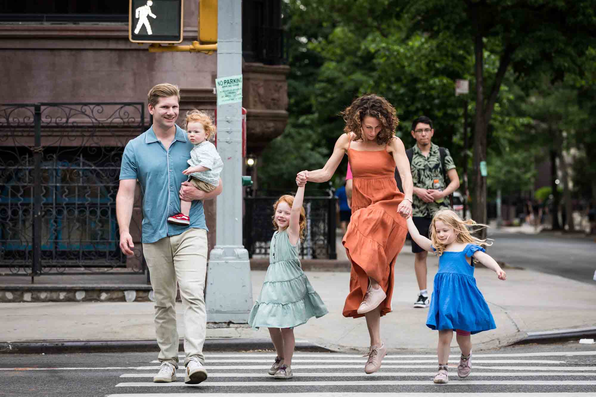 Father holding baby and mother dancing in crosswalk holding hands of two little girls in NYC