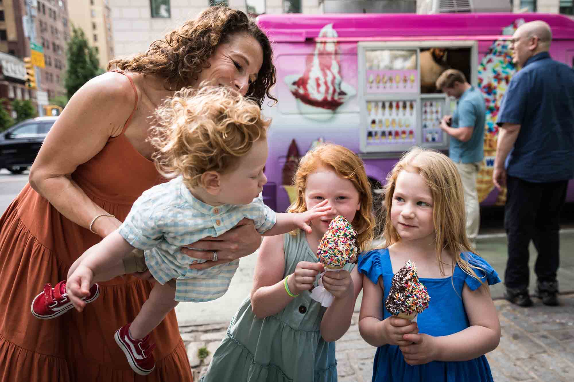 Mother holding baby boy reaching out for colorful ice cream cone held by two little girls on sidewalk