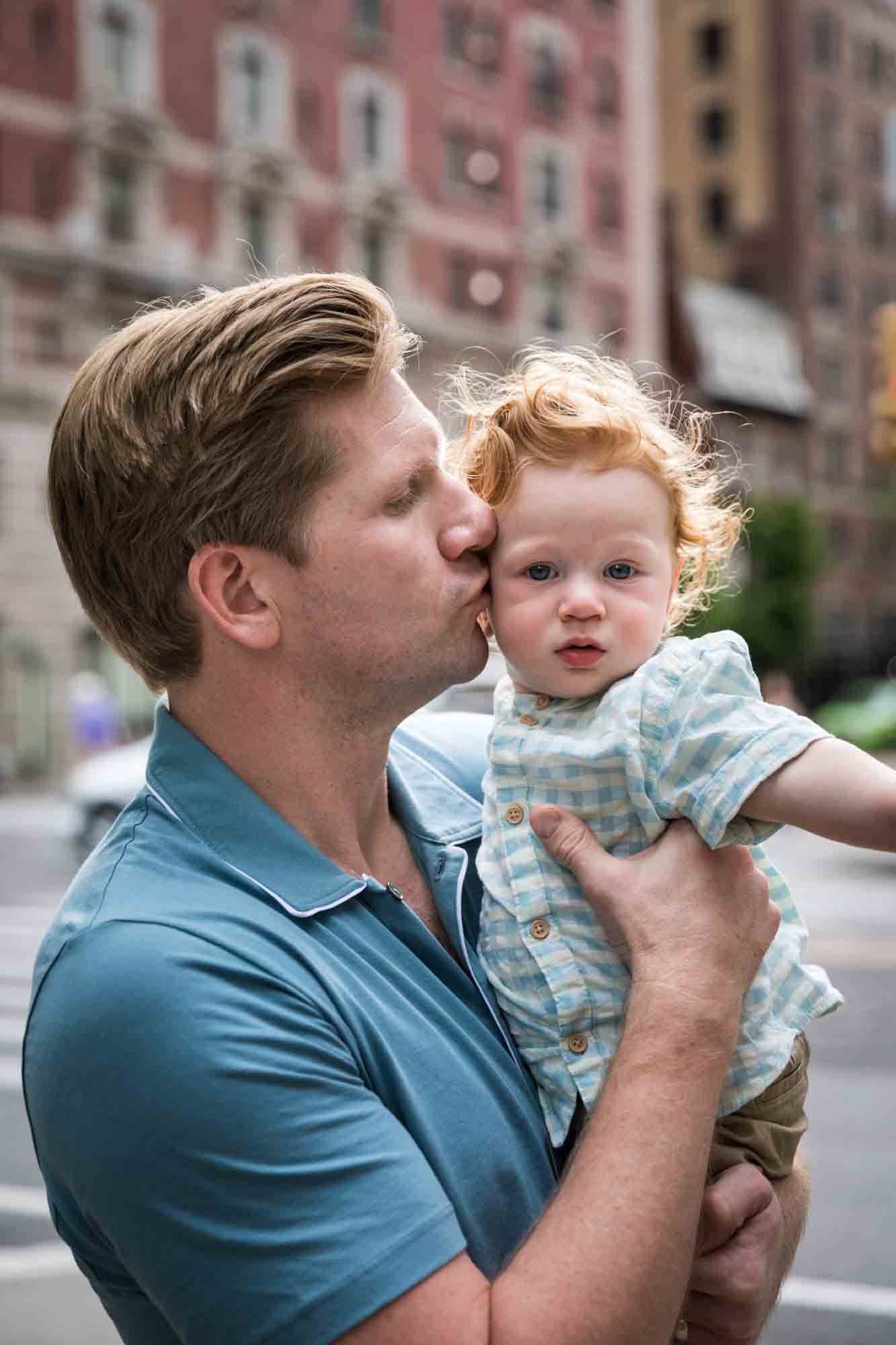 Father wearing blue shirt kissing red-haired baby boy on the side of the head