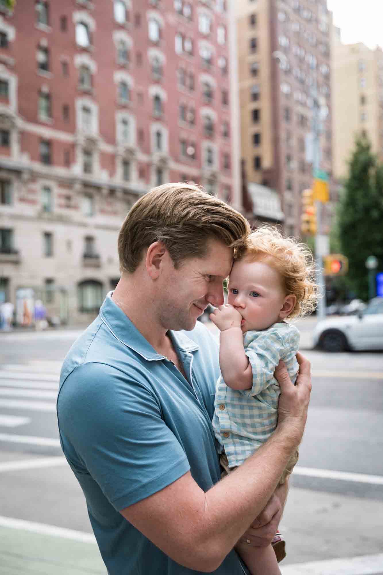 Father wearing blue shirt holding red-haired baby boy in front of NYC street