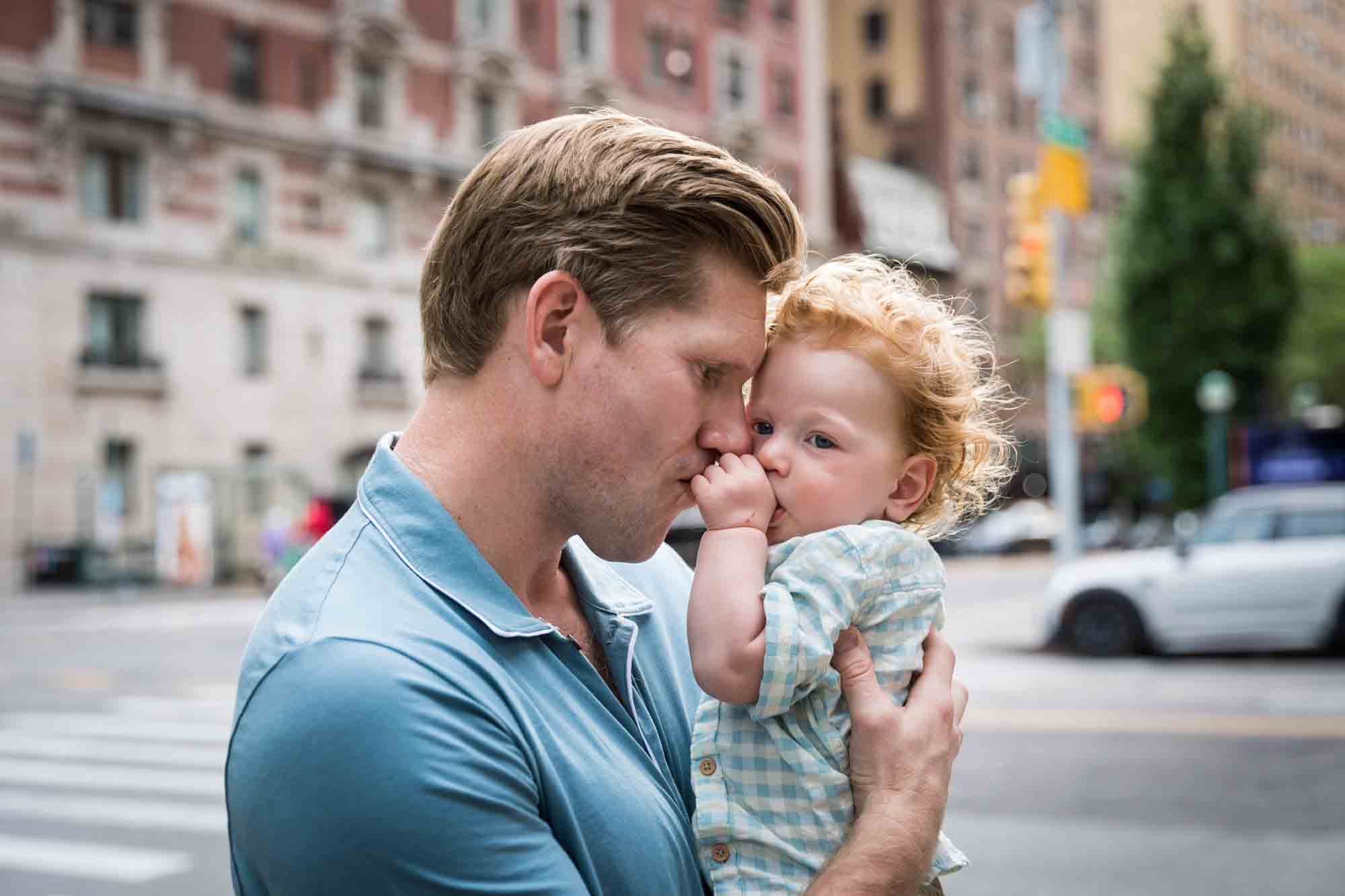 Father wearing blue shirt kissing baby's fist and head pressed up against baby with NYC street in background