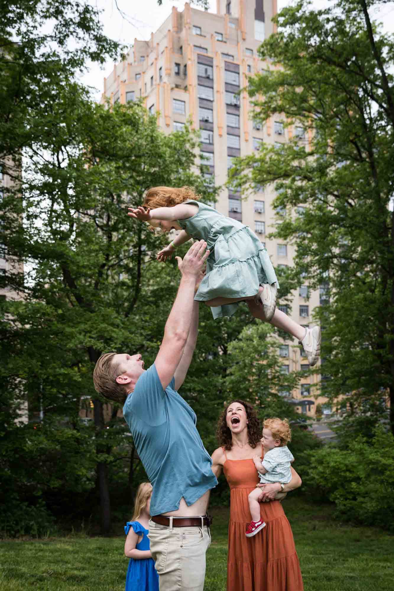 Father throwing little girl in the air with mother reacting in front of building during a Central Park family portrait session