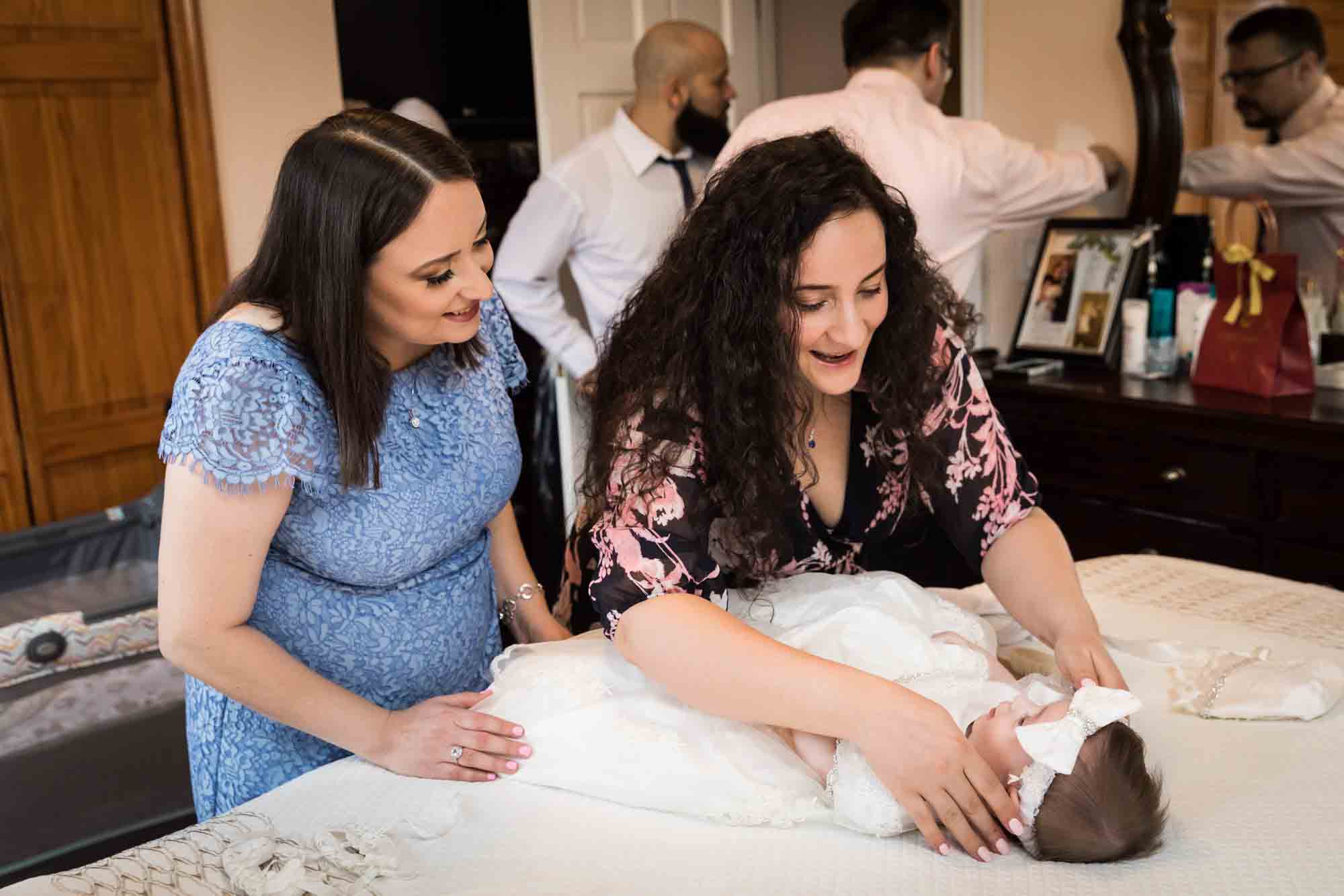 Two female family members dressing baby in white baptism dress in bedroom