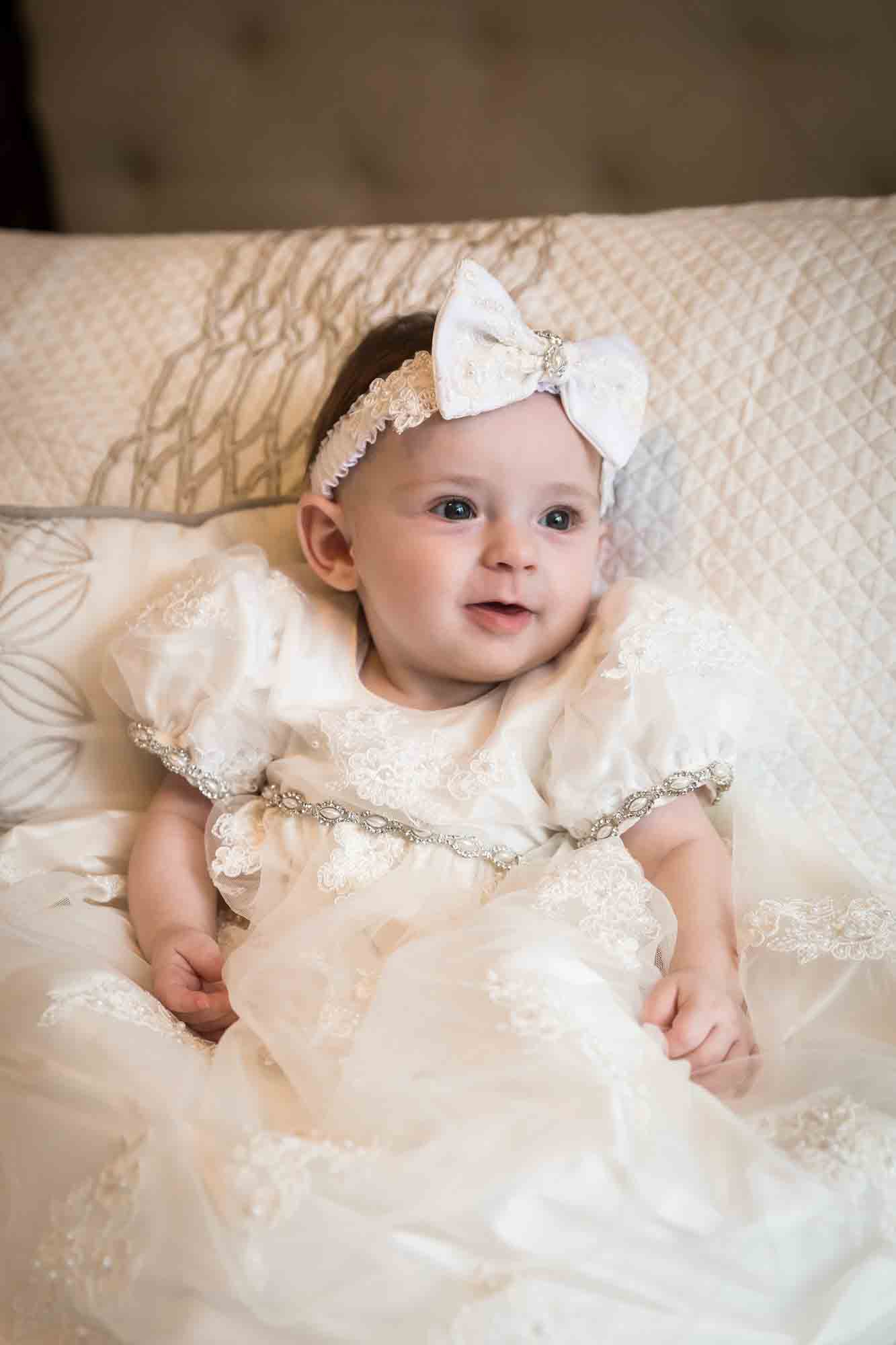Baby girl wearing white baptism dress and white bow sitting in front of pillows