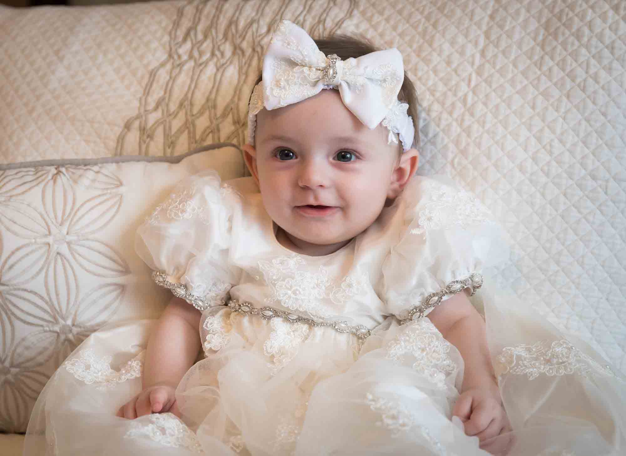 Baby girl wearing white baptism dress and white bow sitting in front of pillows