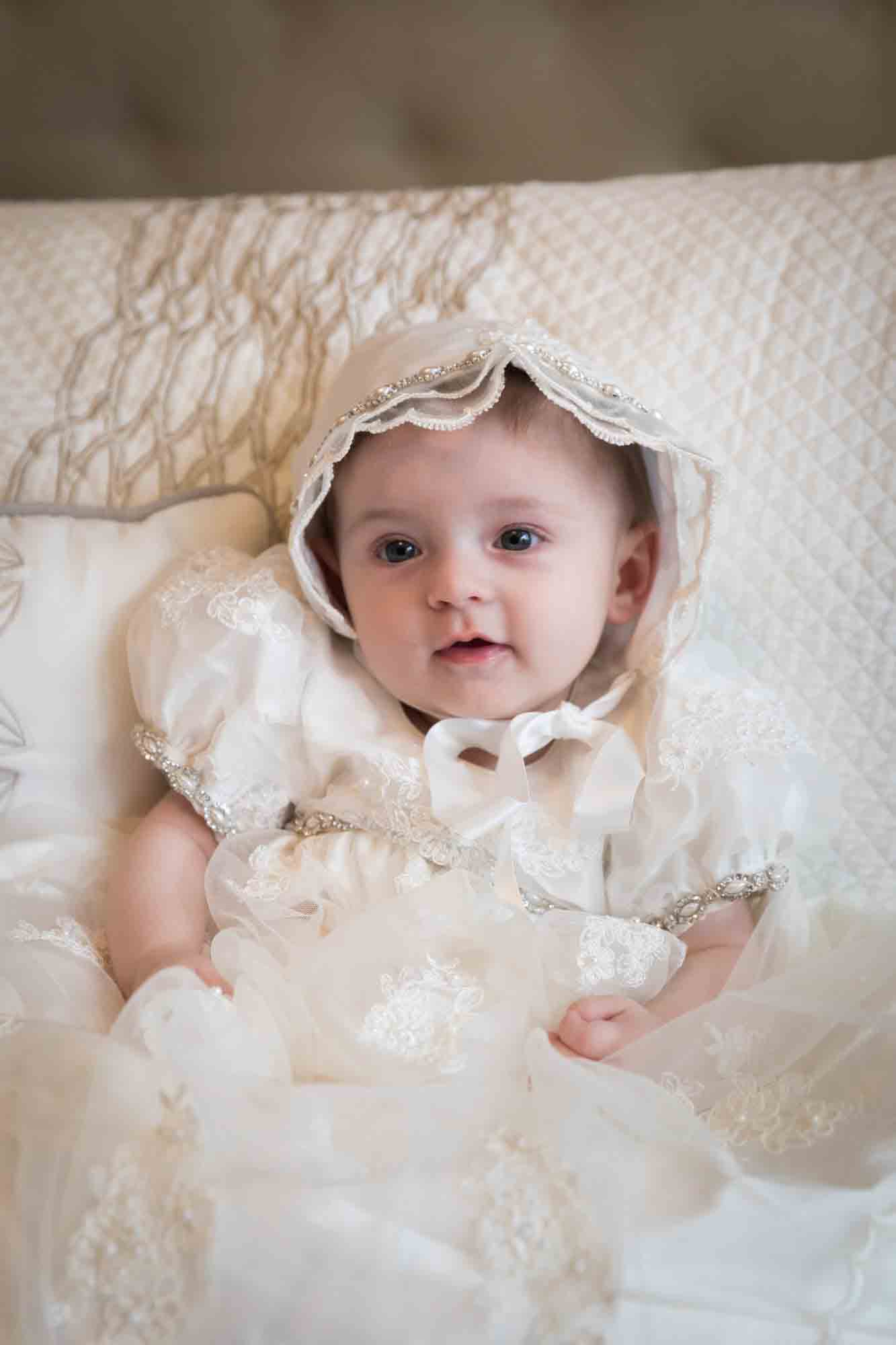 Baby girl wearing white baptism dress and white bonnet sitting in front of pillows
