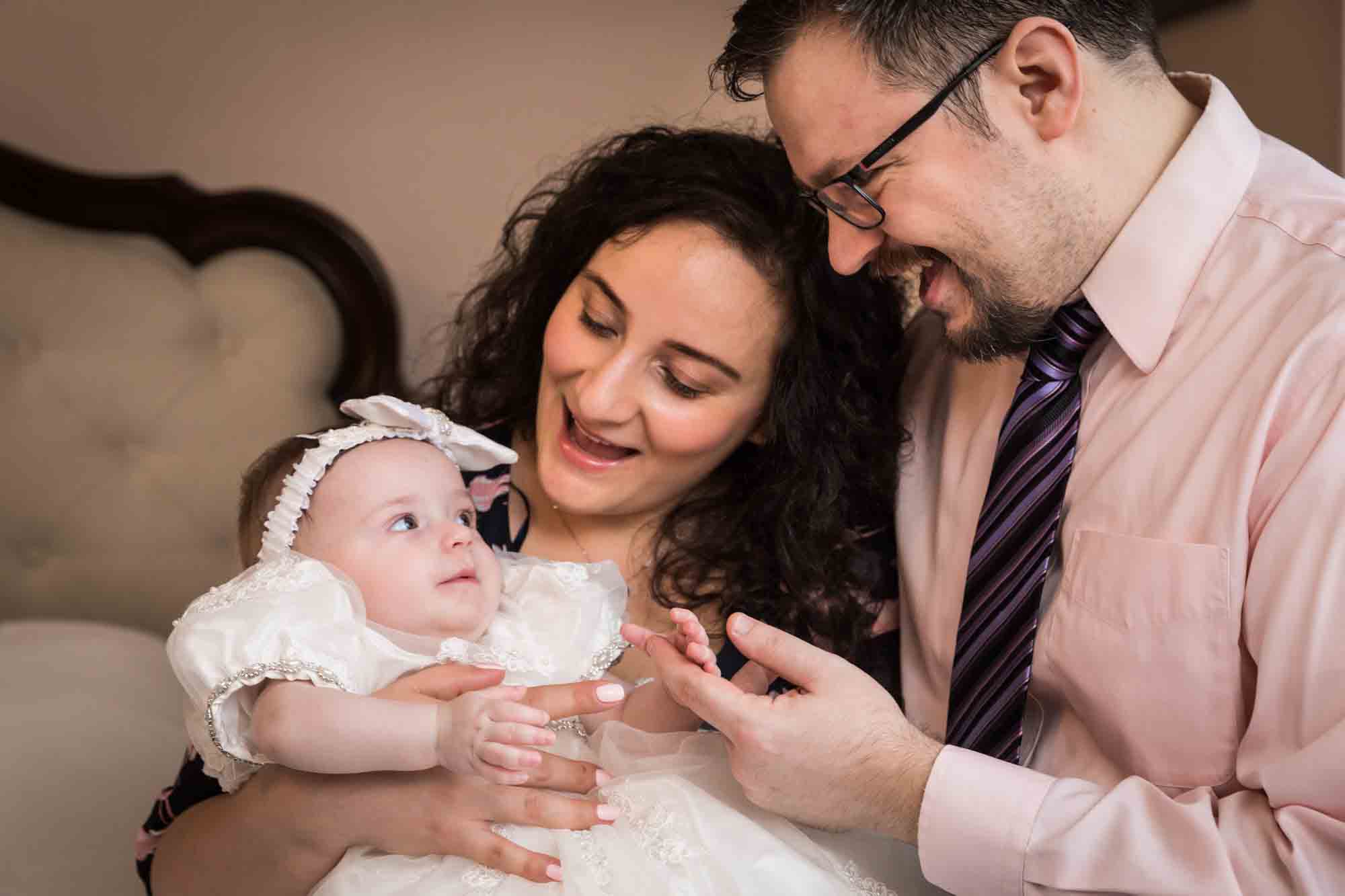 Mother and father playing with baby girl wearing white baptism dress and hair bow