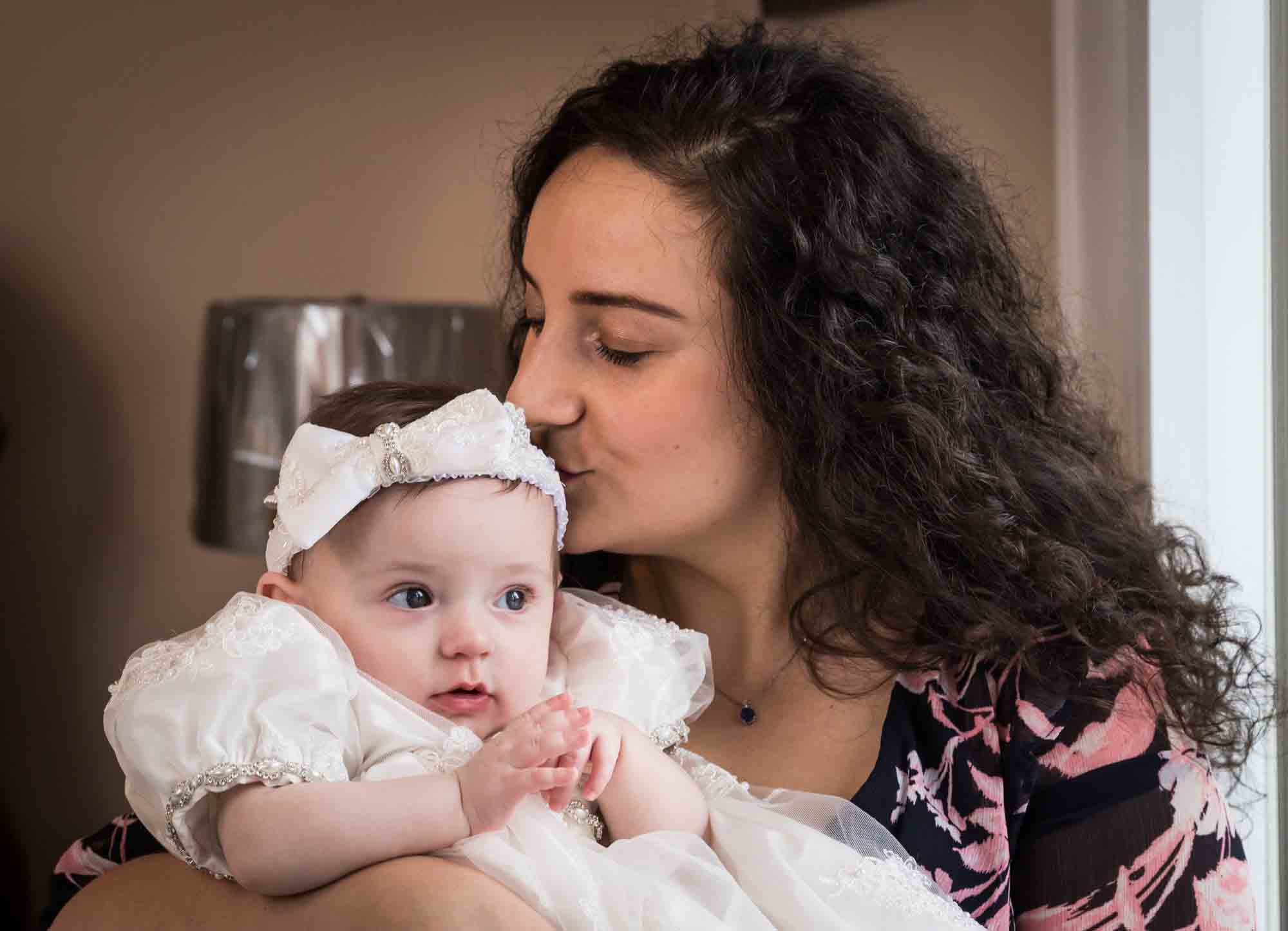 Mother kissing baby girl wearing white baptism dress and hair bow