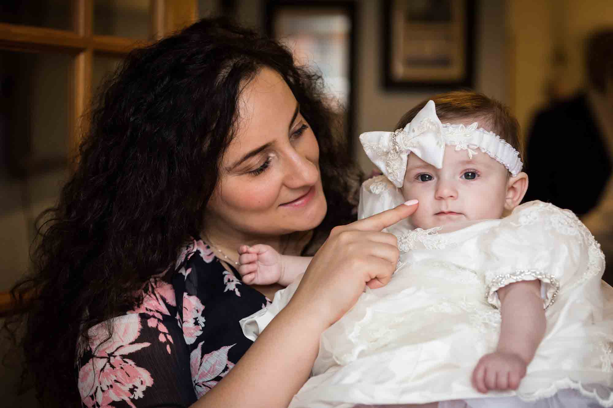 Mother touching nose of baby girl wearing white baptism dress and hair bow