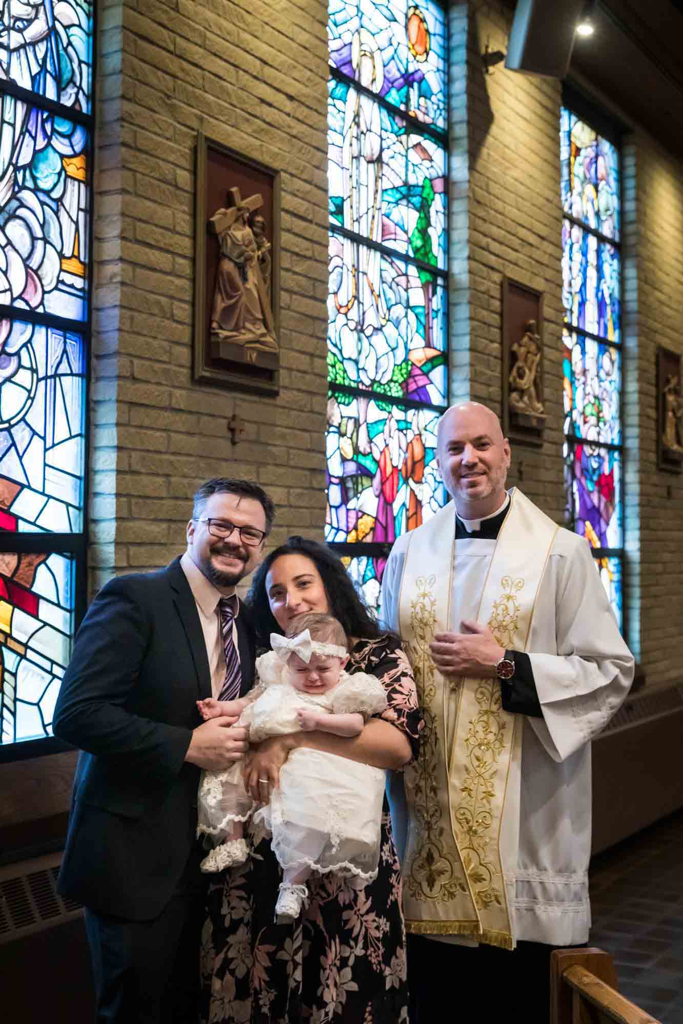 Maspeth baptism photos of family holding baby with priest in front of stained glass windows