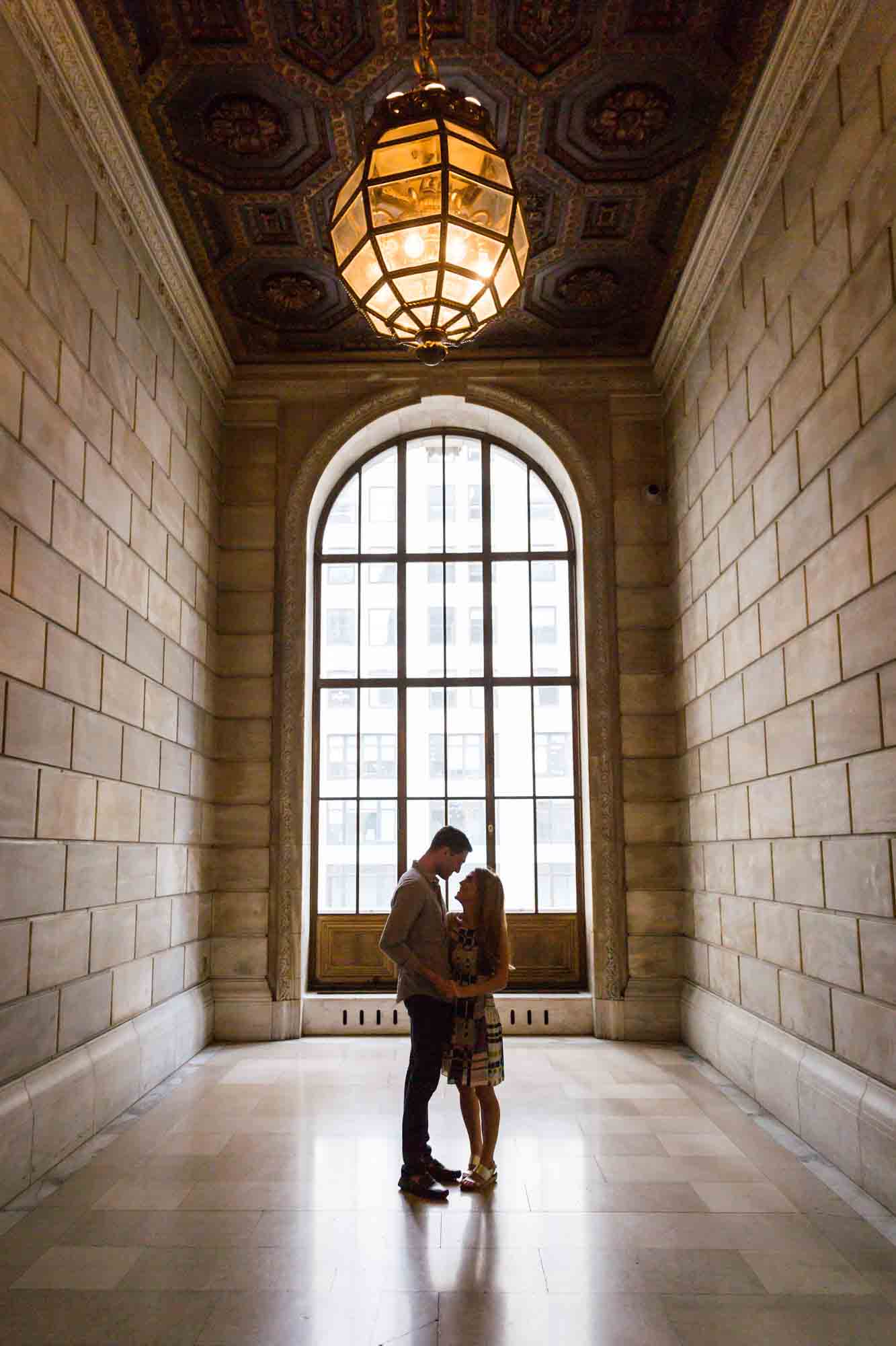 Couple holding hands together in front of a large rounded window under a round chandelier during a New York public library engagement photo shoot