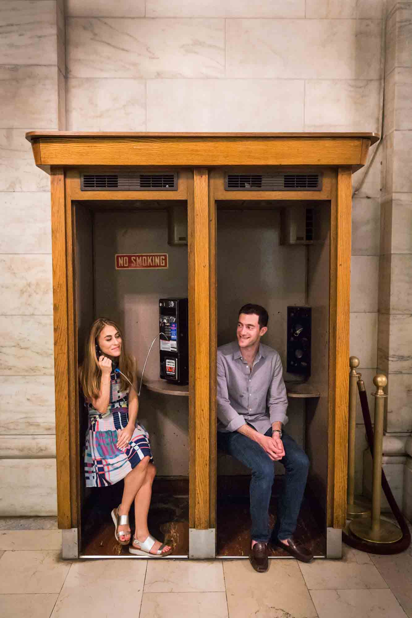 Couple sitting at a vintage phone booth in a marble hall during a New York public library engagement photo shoot