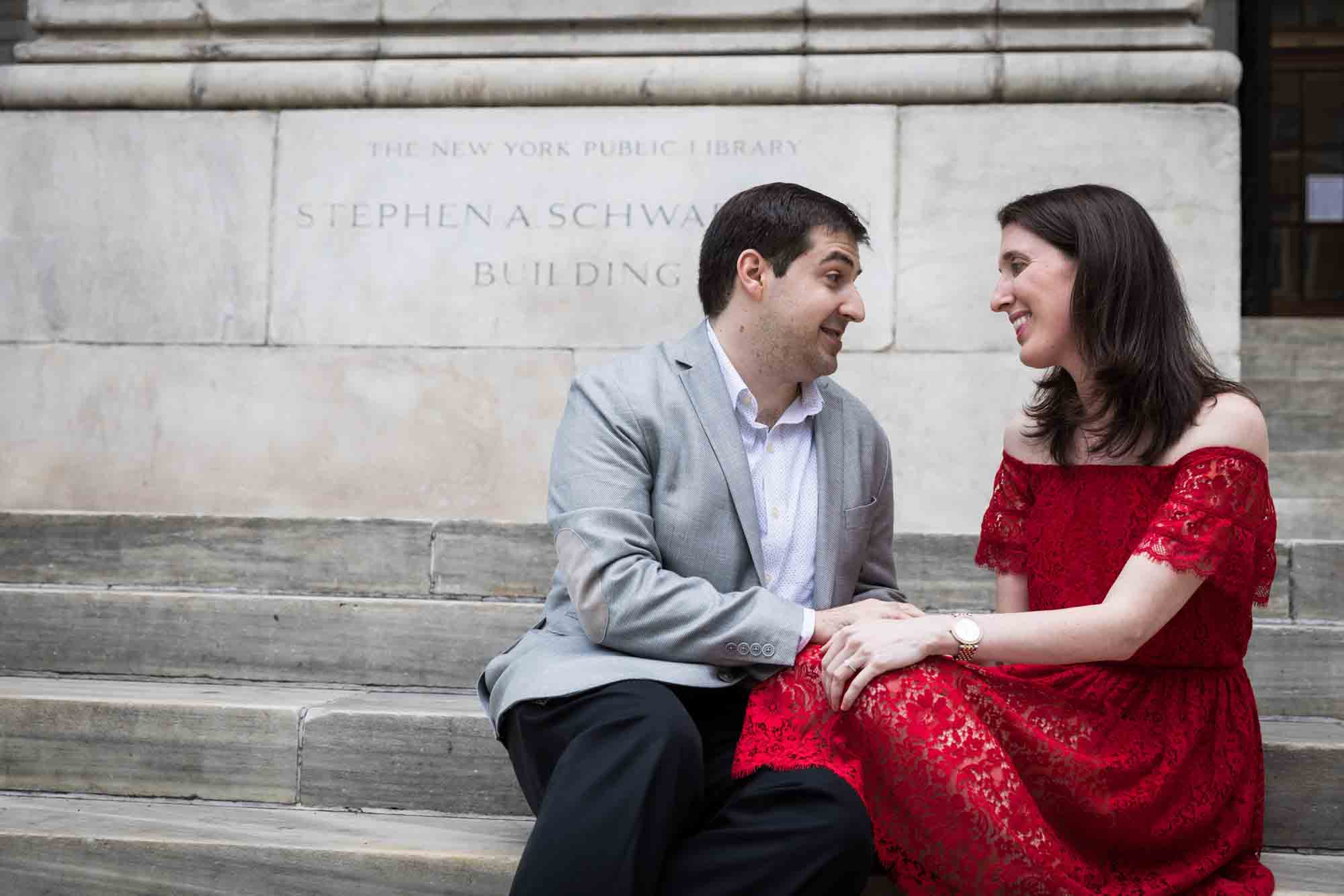 Woman in red dress holding hands of man in grey jacket in front of stone sign during New York Public Library proposal