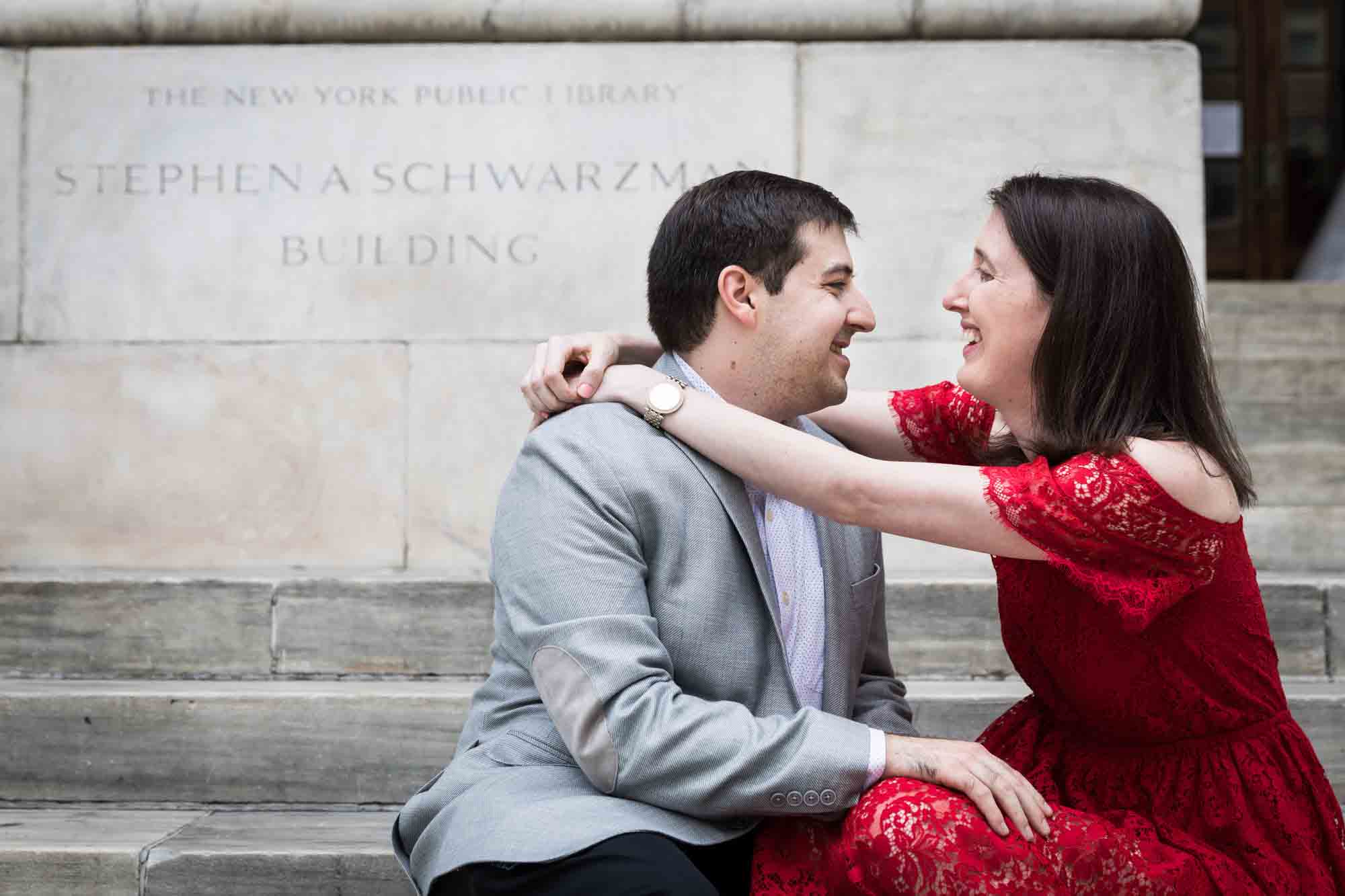 Woman in red dress hugging man in grey jacket in front of stone sign during New York Public Library proposal