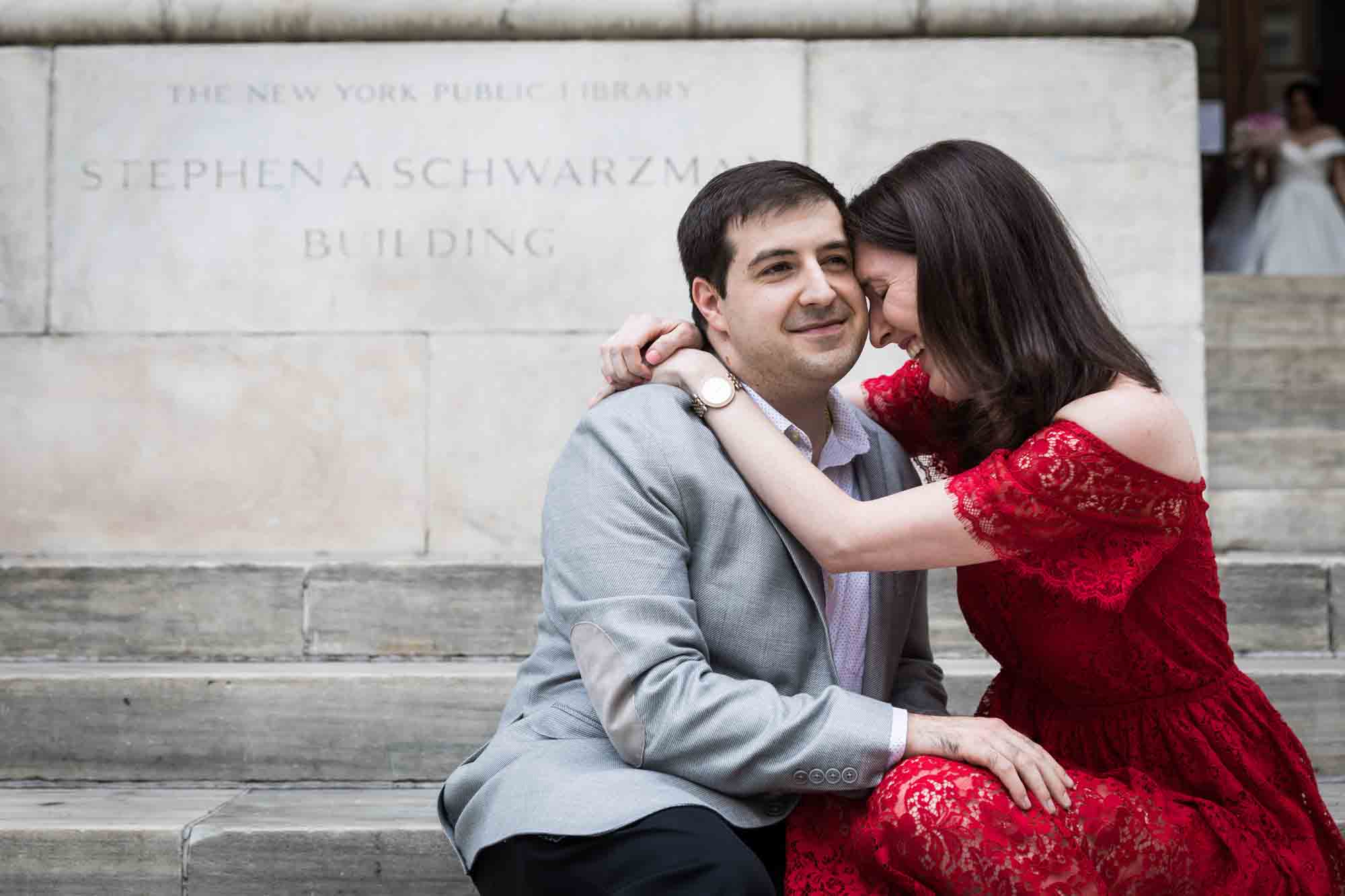 Woman in red dress hugging man in grey jacket in front of stone sign during New York Public Library proposal