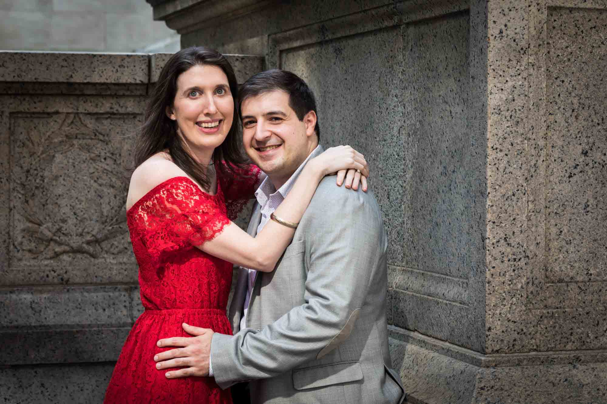Man hugging in front of marble alcove with woman wearing red dress during a New York Public Library surprise proposal