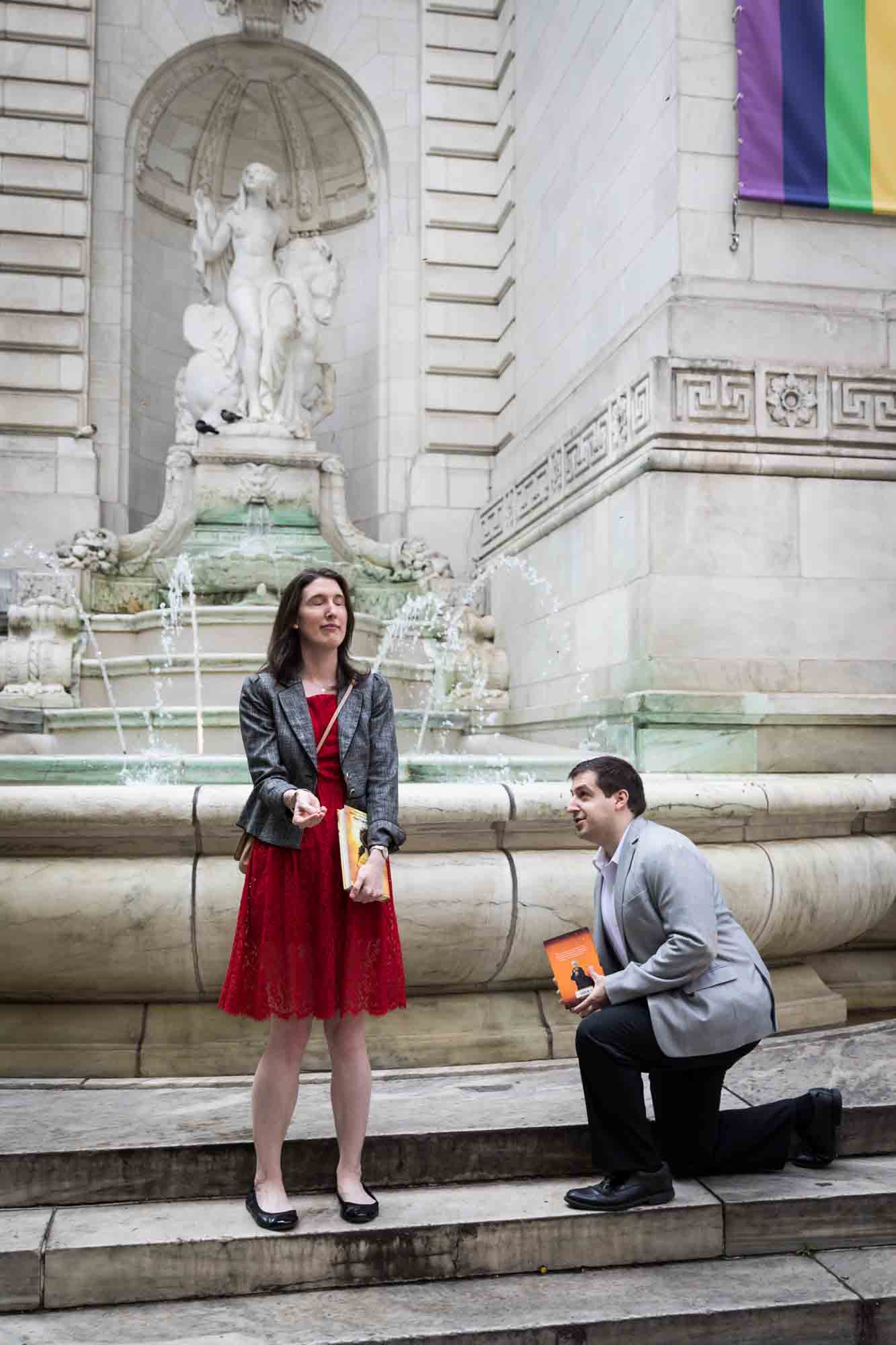 Woman wearing red dress and man on one knee in front of 'Beauty' fountain during a New York Public Library surprise proposal