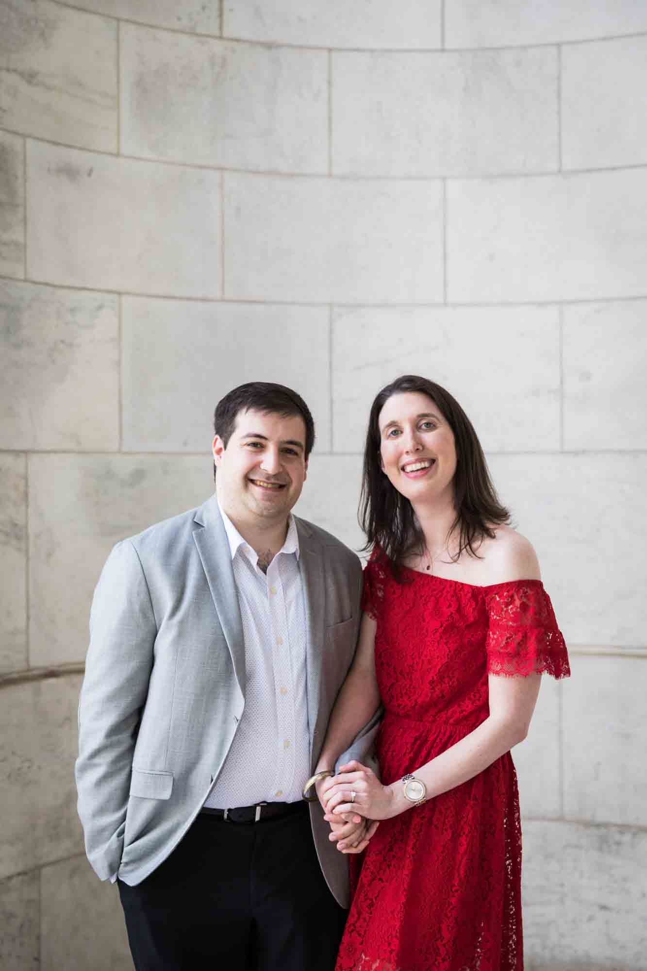 Man holding hands in marble alcove with woman wearing red dress during a New York Public Library surprise proposal