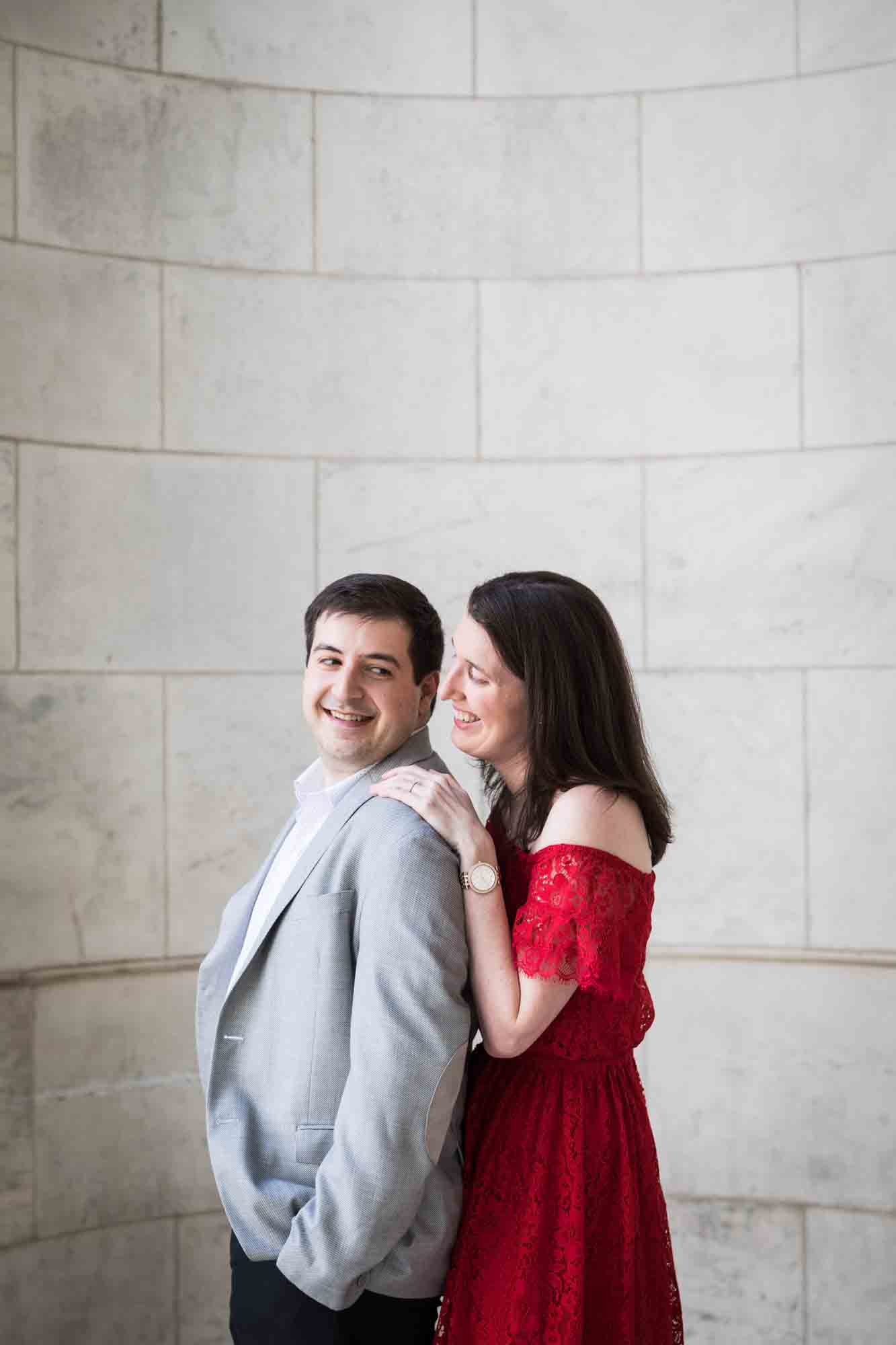 Man hugging in marble alcove with woman wearing red dress during a New York Public Library surprise proposal