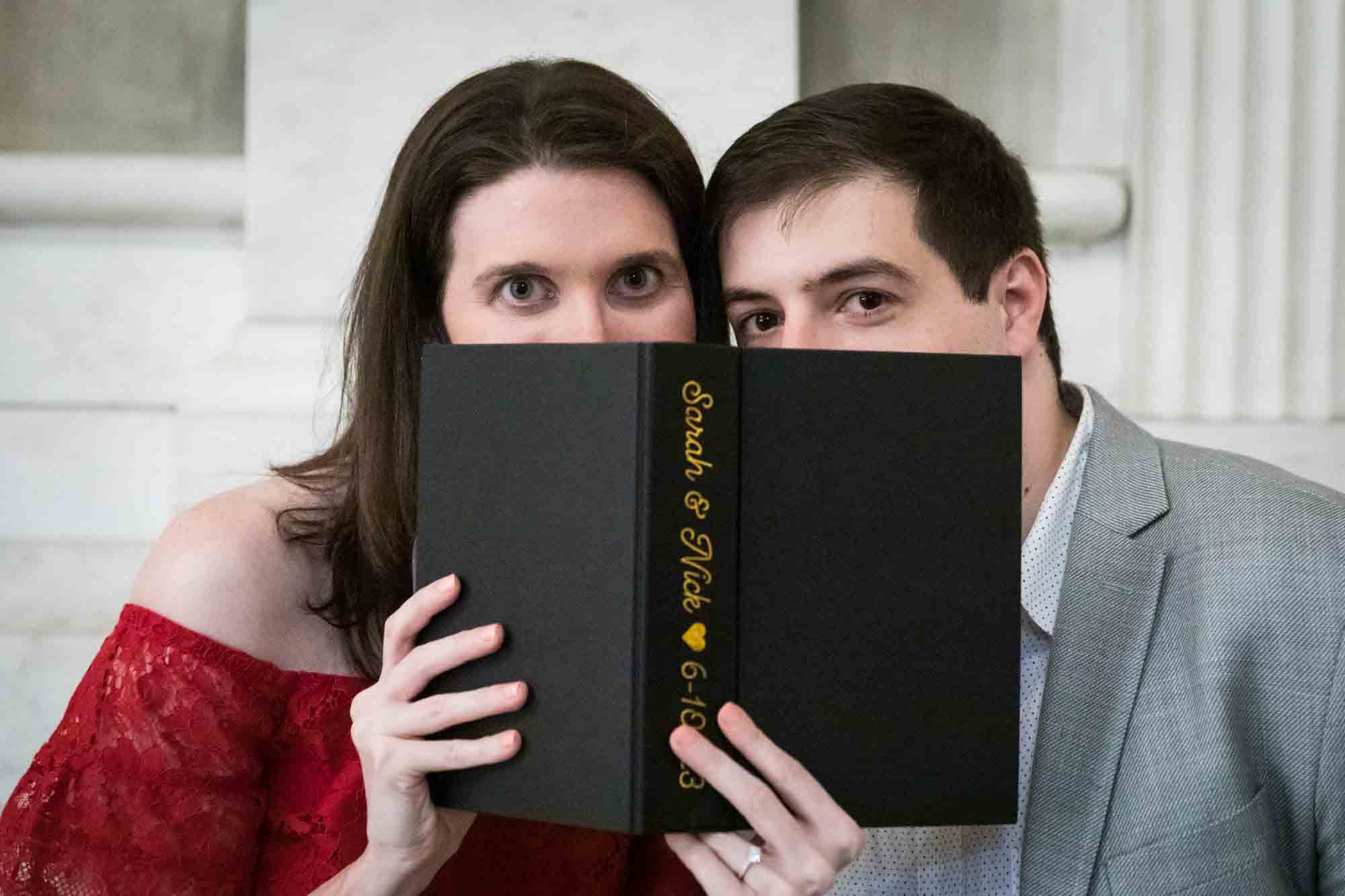 Couple peeking over black book with woman wearing red dress during a New York Public Library surprise proposal