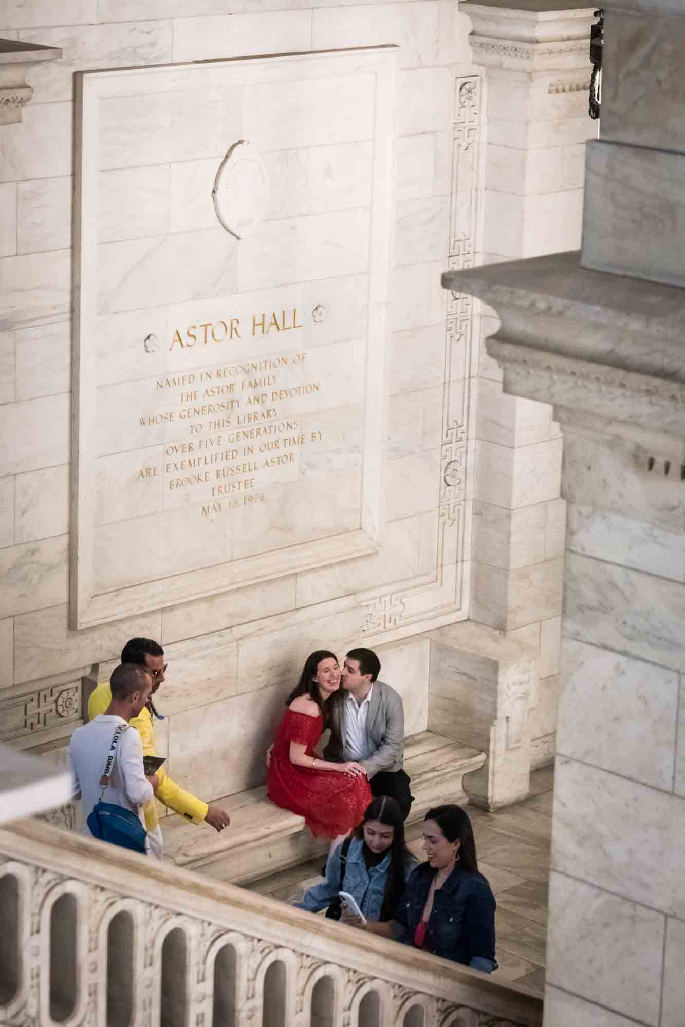 Couple kissing on stone bench in staircase with patrons walking past during a New York Public Library surprise proposal