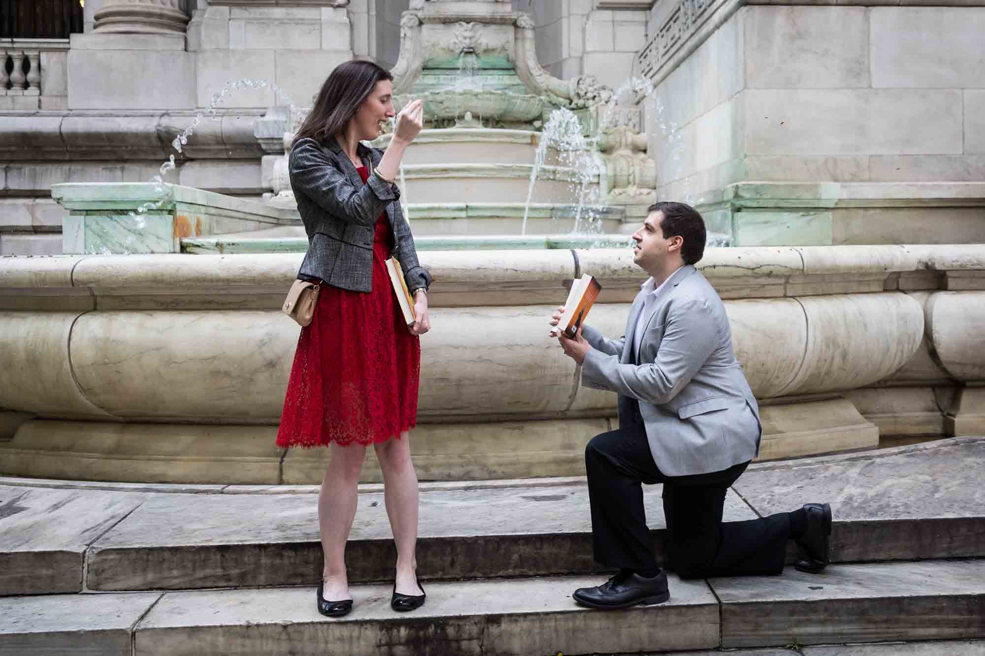 Woman in red dress about to cry in front of man wearing grey jacket on one knee holding book during New York Public Library proposal