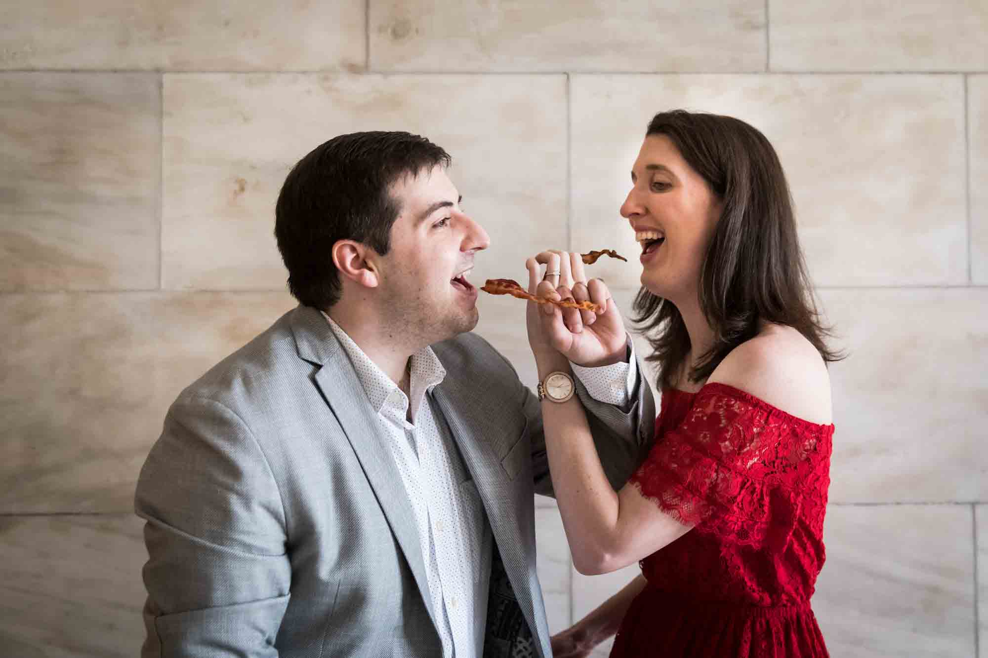 Couple feeding each other bacon in front of marble wall during a New York Public Library surprise proposal