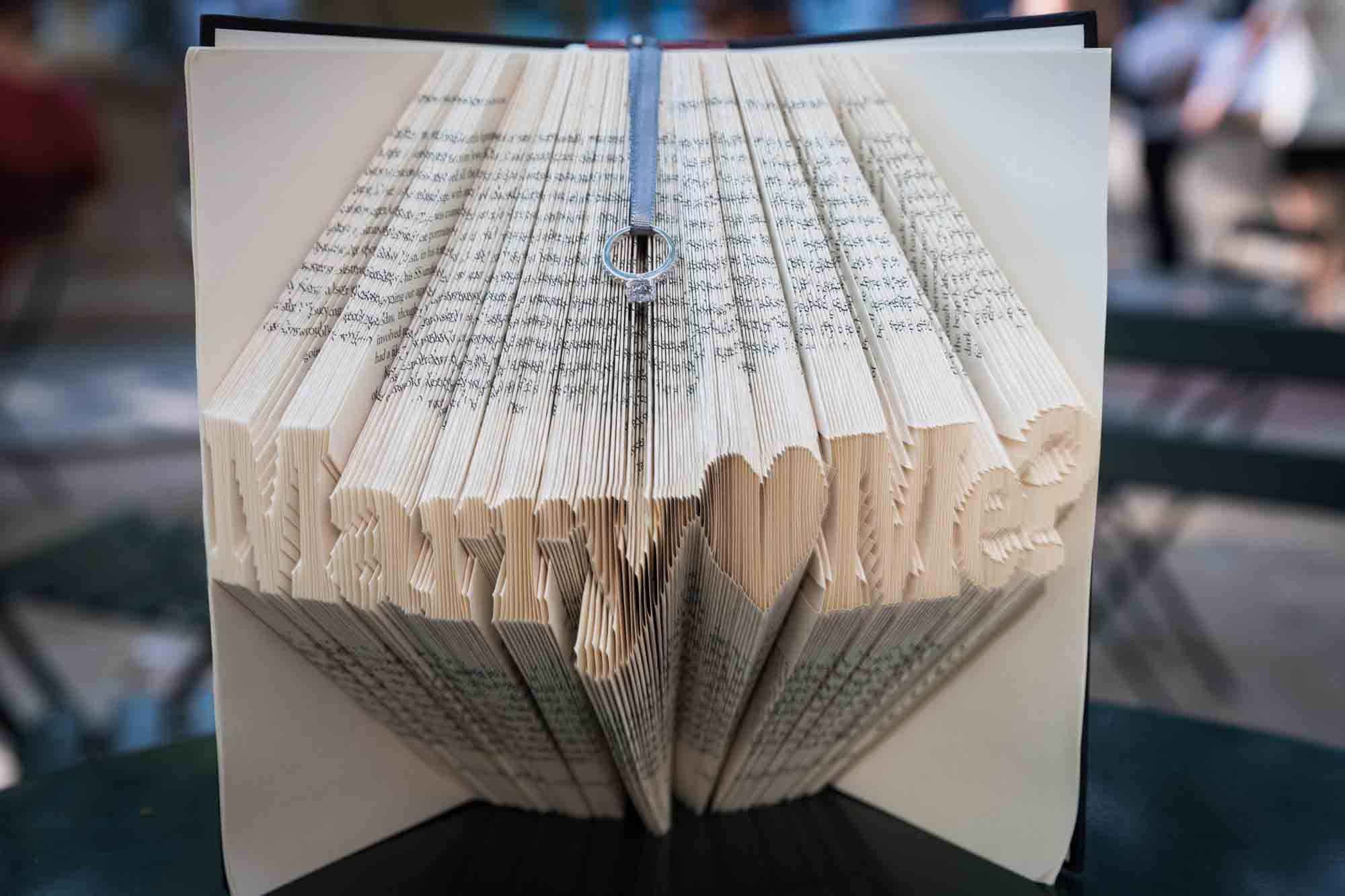 Intricately folded book with the words 'Marry Me?' formed and engagement ring held by ribbon on top