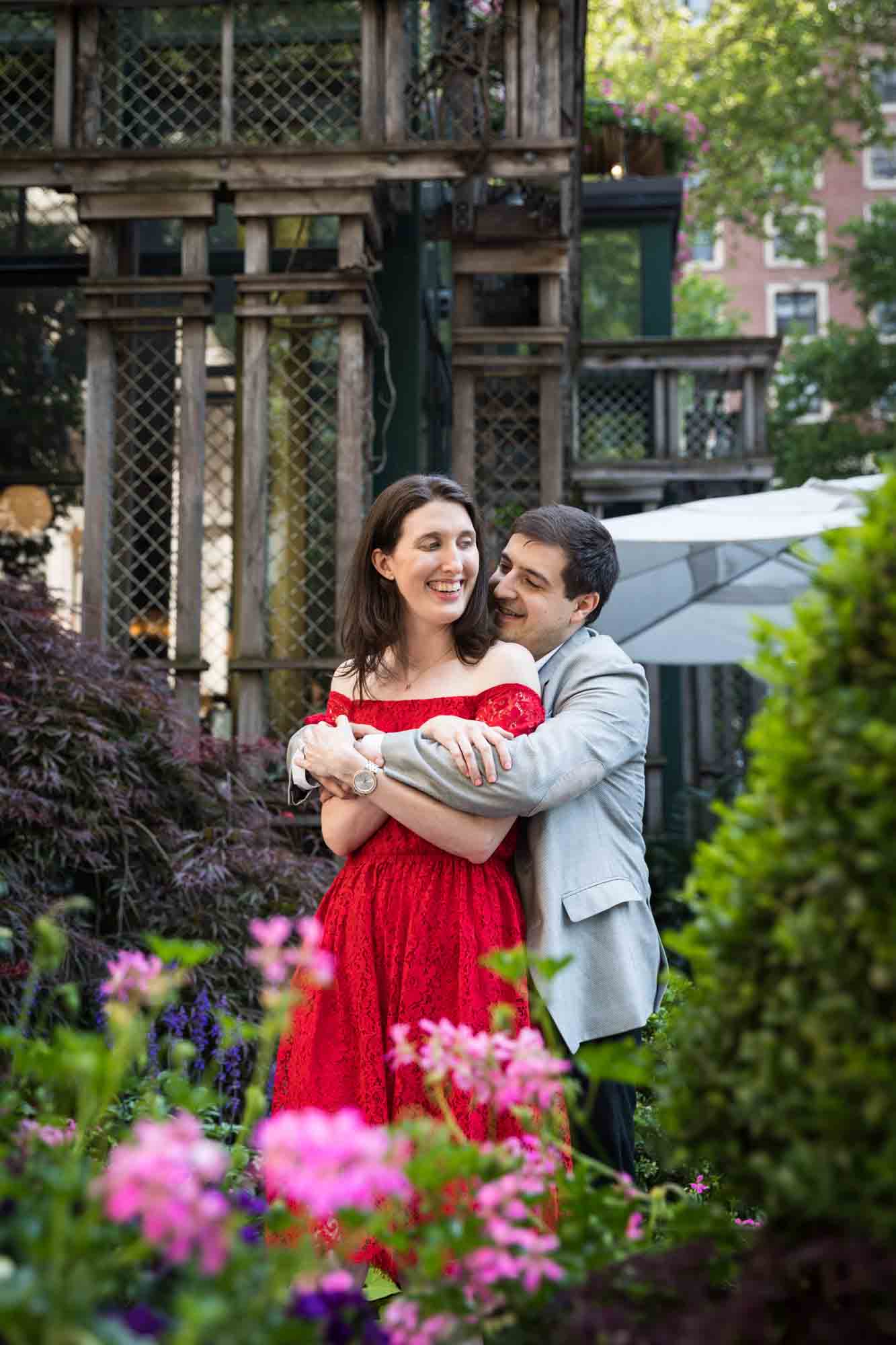 Man in grey jacket hugging woman in red dress in Bryant Park behind pink flowers