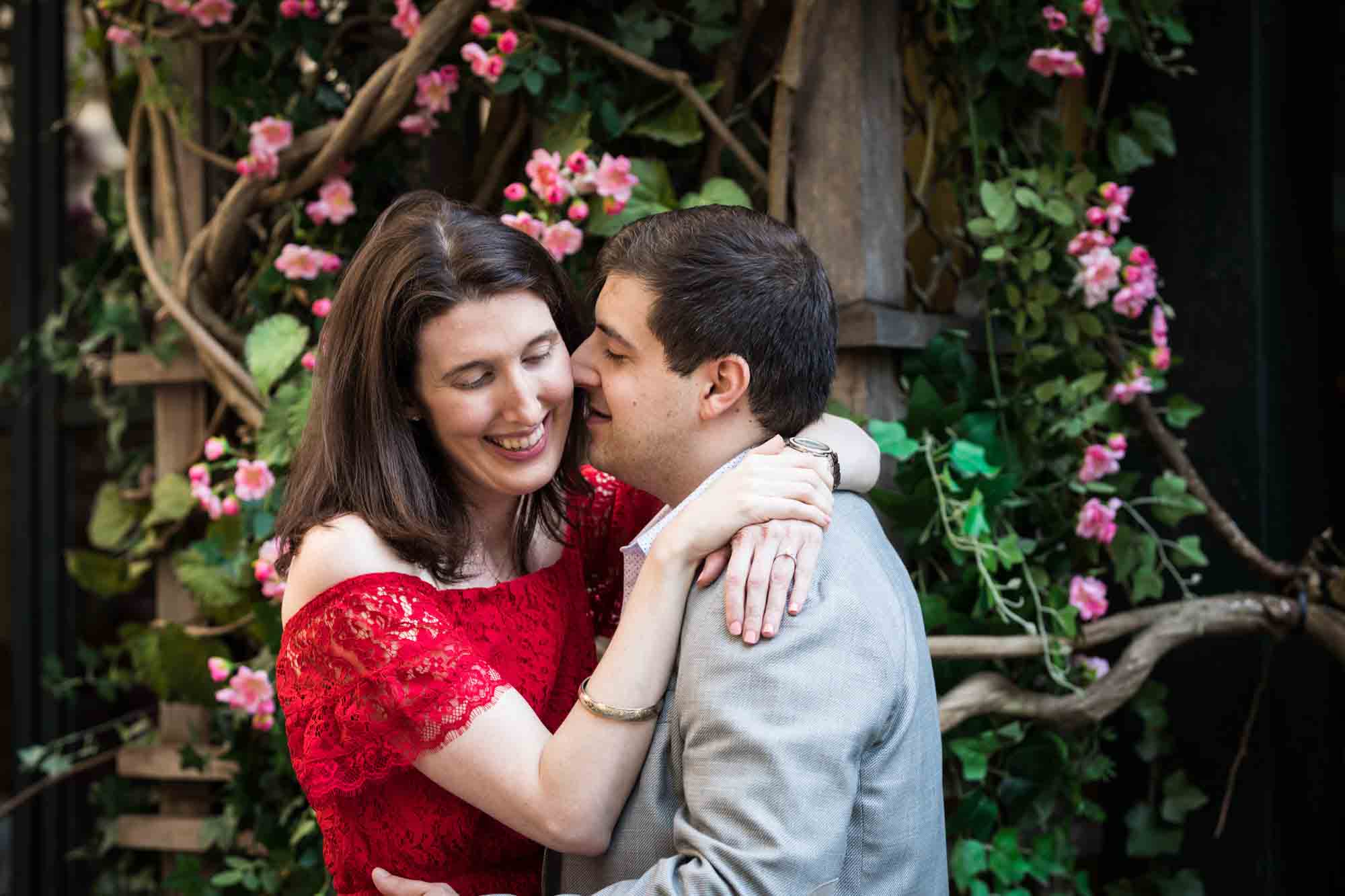 Man in grey jacket hugging woman in red dress in Bryant Park in front of pink flowers