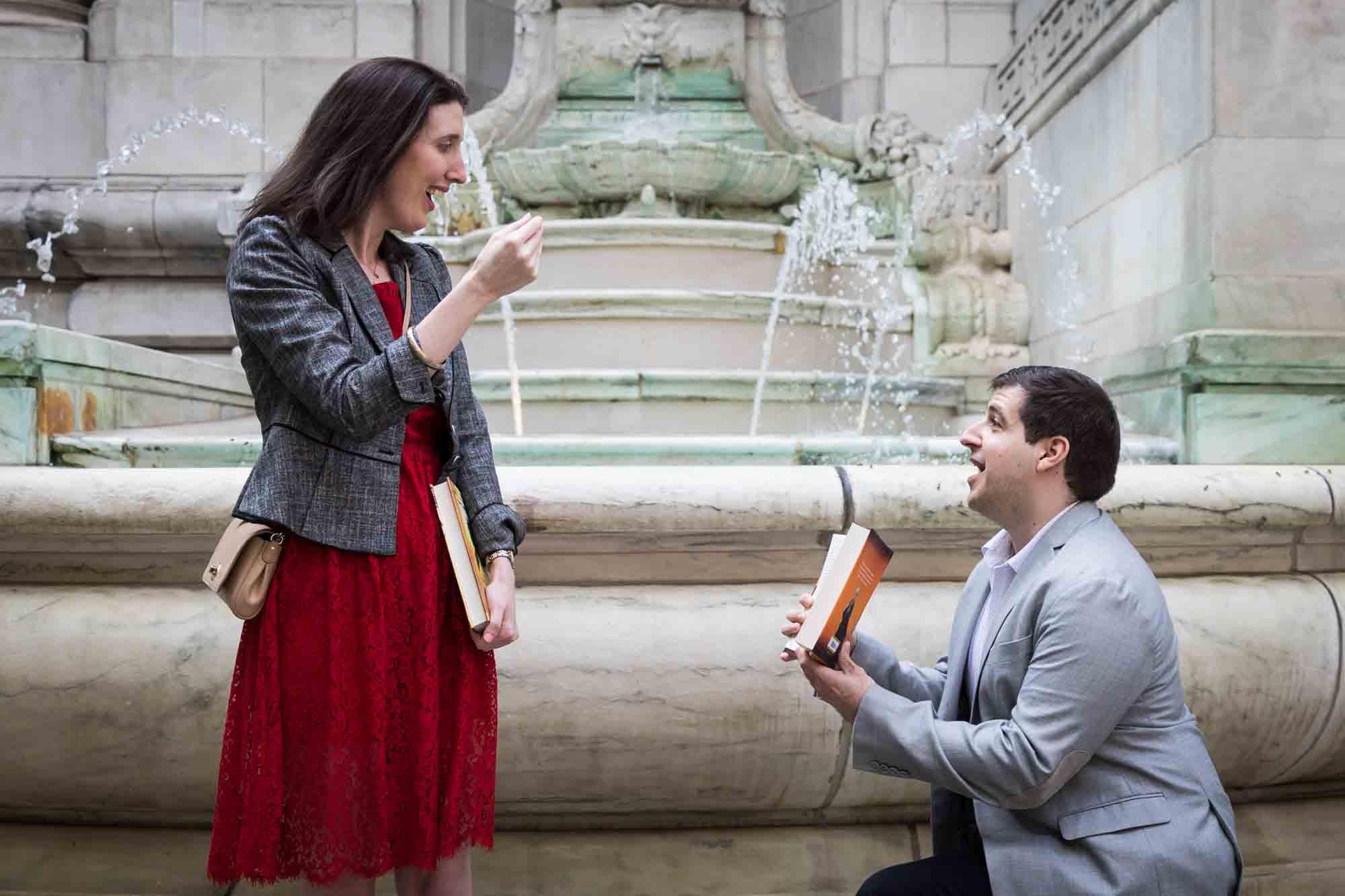 Woman in red dress in front of man wearing grey jacket on one knee holding book during New York Public Library proposal