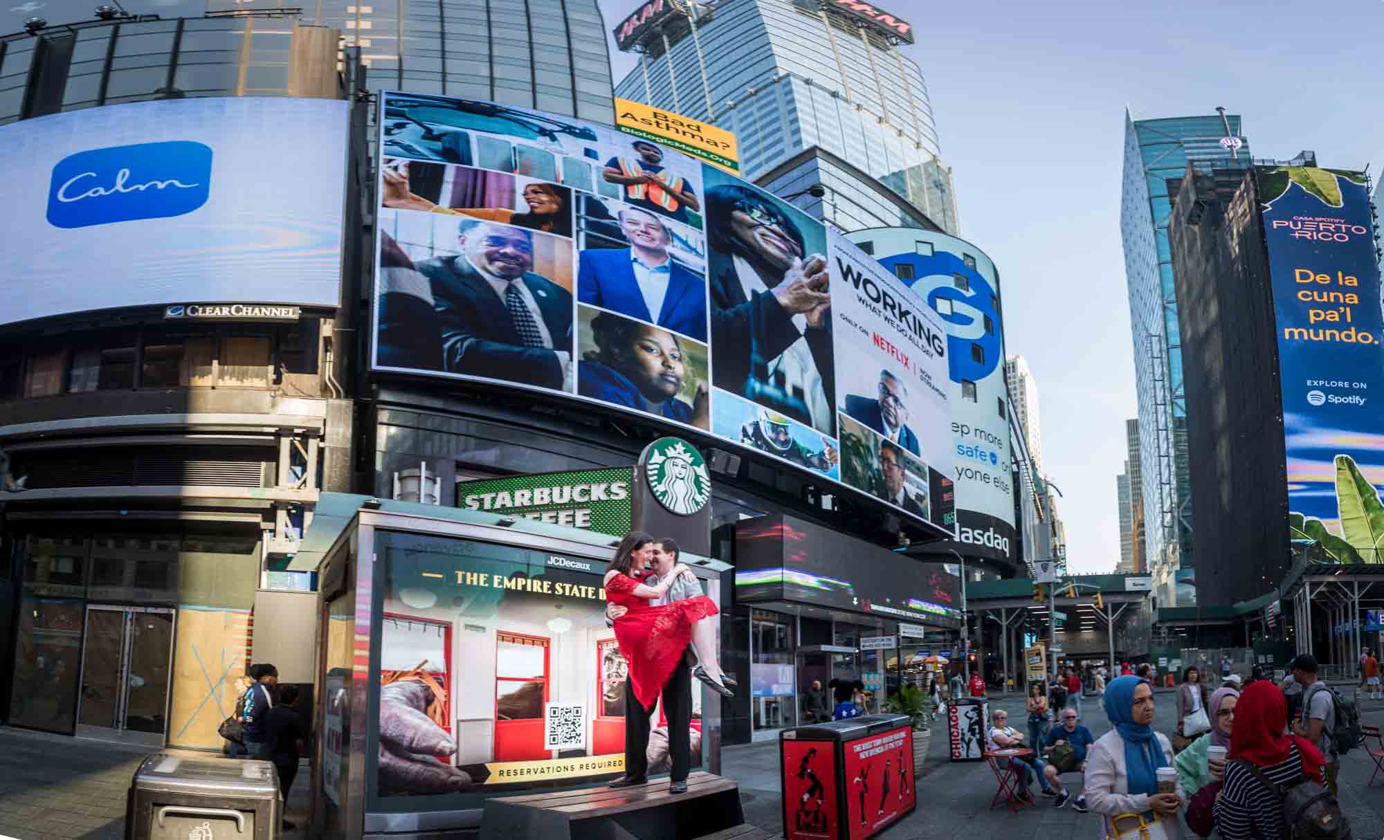 Man holding woman wearing red dress on platform in Times Square in front of colorful billboards
