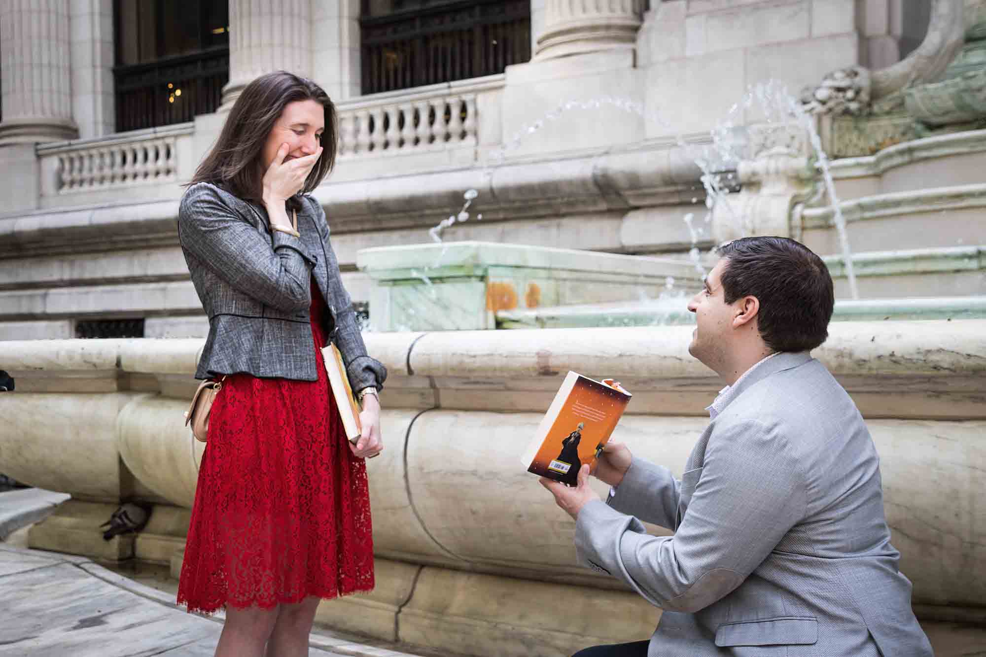 Woman in red dress about to cry in front of man wearing grey jacket on one knee holding book during New York Public Library proposal