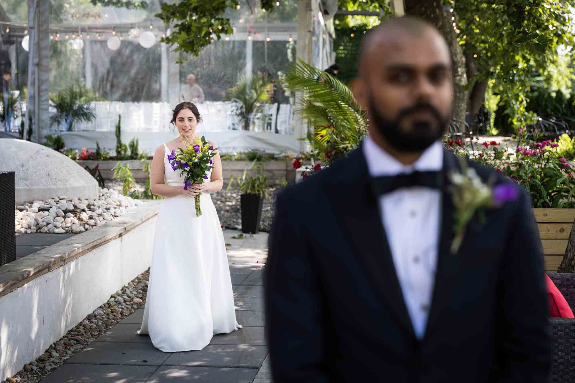 Bride wearing white dress and holding bouquet looking at groom wearing tux in foreground during first look at a Sanctuary Roosevelt Island wedding