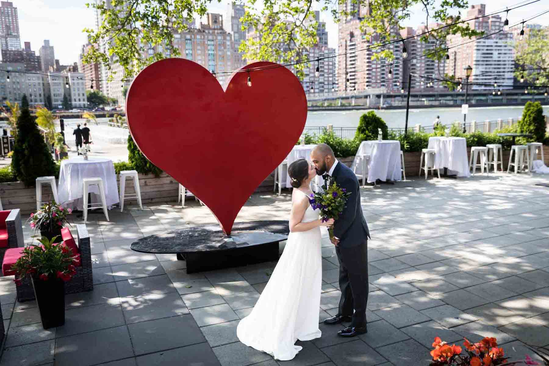 Bride and groom kissing on patio in front of red heart sculpture at a Sanctuary Roosevelt Island wedding