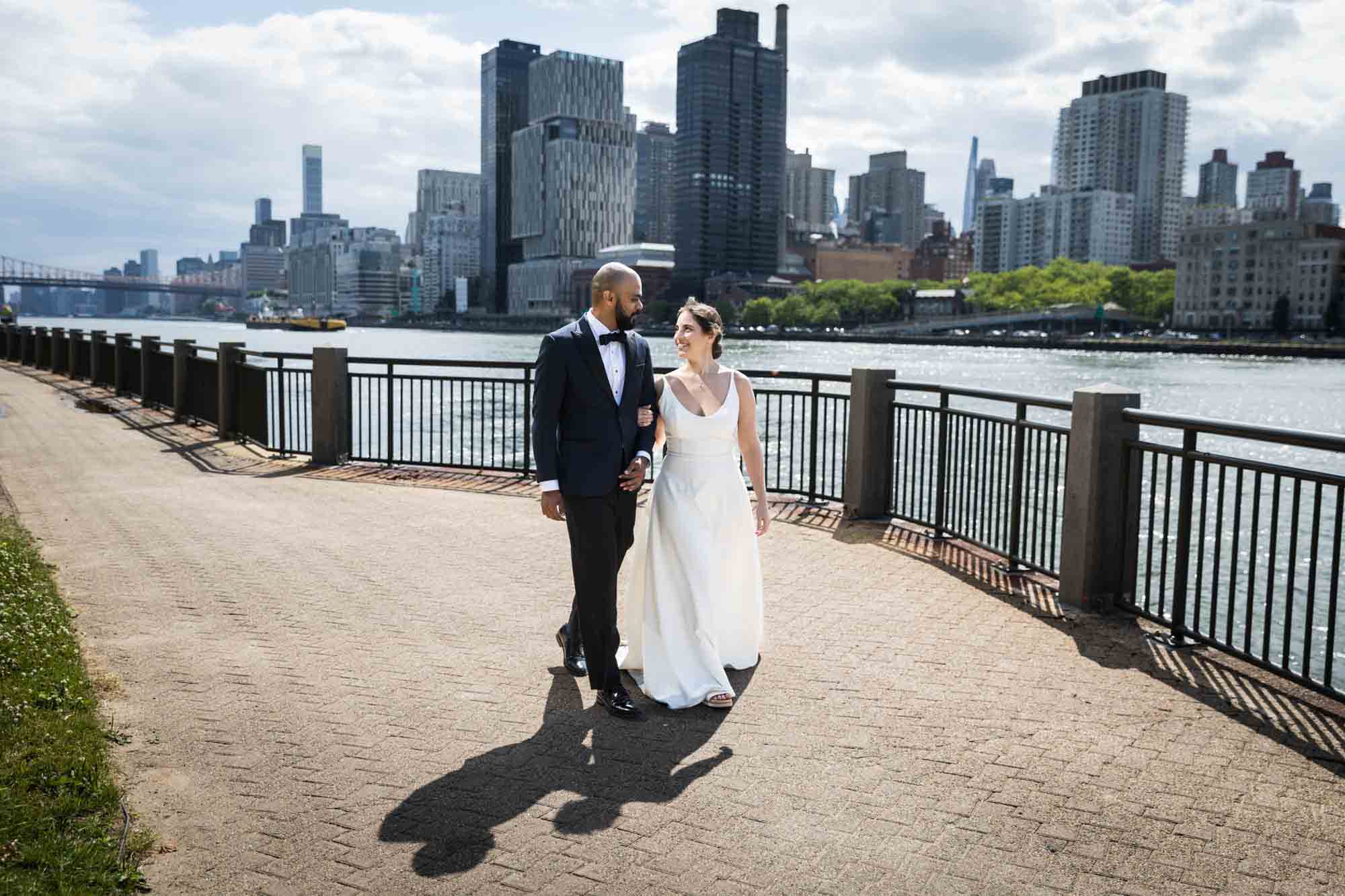 Bride and groom walking along waterfront in front of NYC skyline on Roosevelt Island